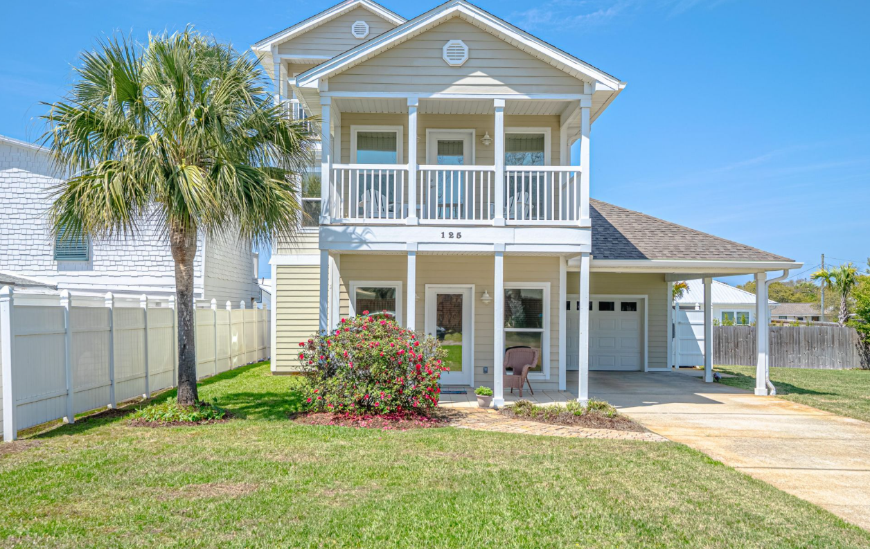 A two-story tan coastal-style house with a front porch, a balcony, and a palm tree in the front yard.
