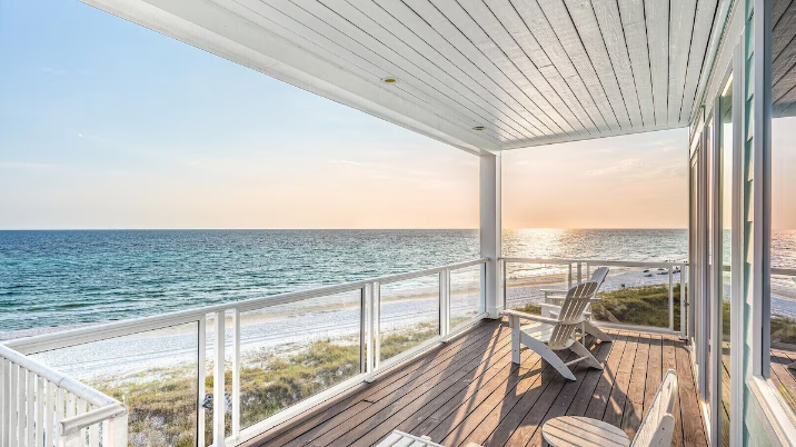 A wooden deck with a white railing overlooking a sandy beach and the ocean during a golden sunset.