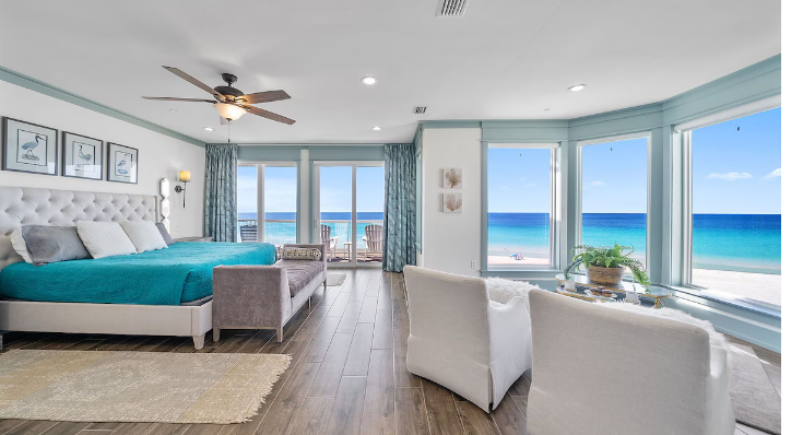 A modern master bedroom with a large bed, seating area, and floor-to-ceiling windows overlooking a turquoise ocean.