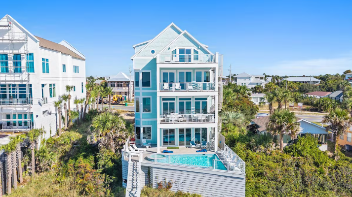 A multi-story, light-blue house featuring balconies and an outdoor pool, surrounded by greenery under a clear blue sky.