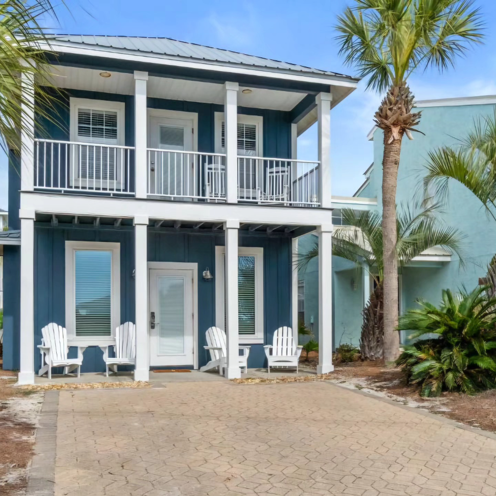 Two-story dark blue house with white trim, a front porch, a balcony, white Adirondack chairs, and a palm tree.
