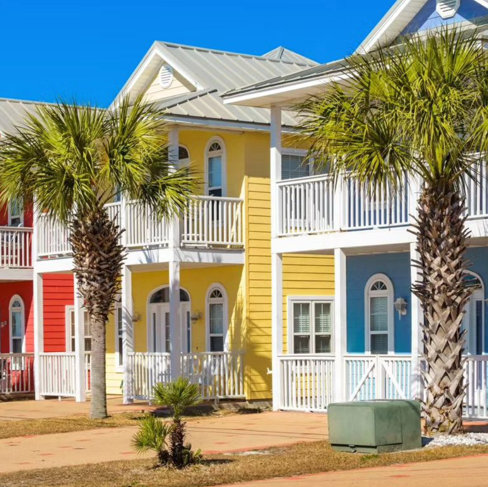 A row of vibrant, multi-colored coastal houses with white balconies and palm trees under a bright blue sky.