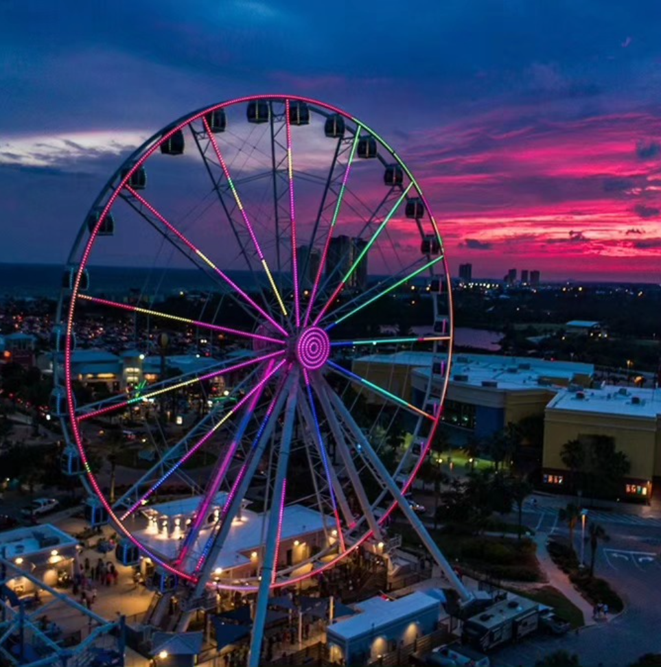 A colorful, illuminated Ferris wheel stands against a vibrant pink and blue sunset over a coastal town.