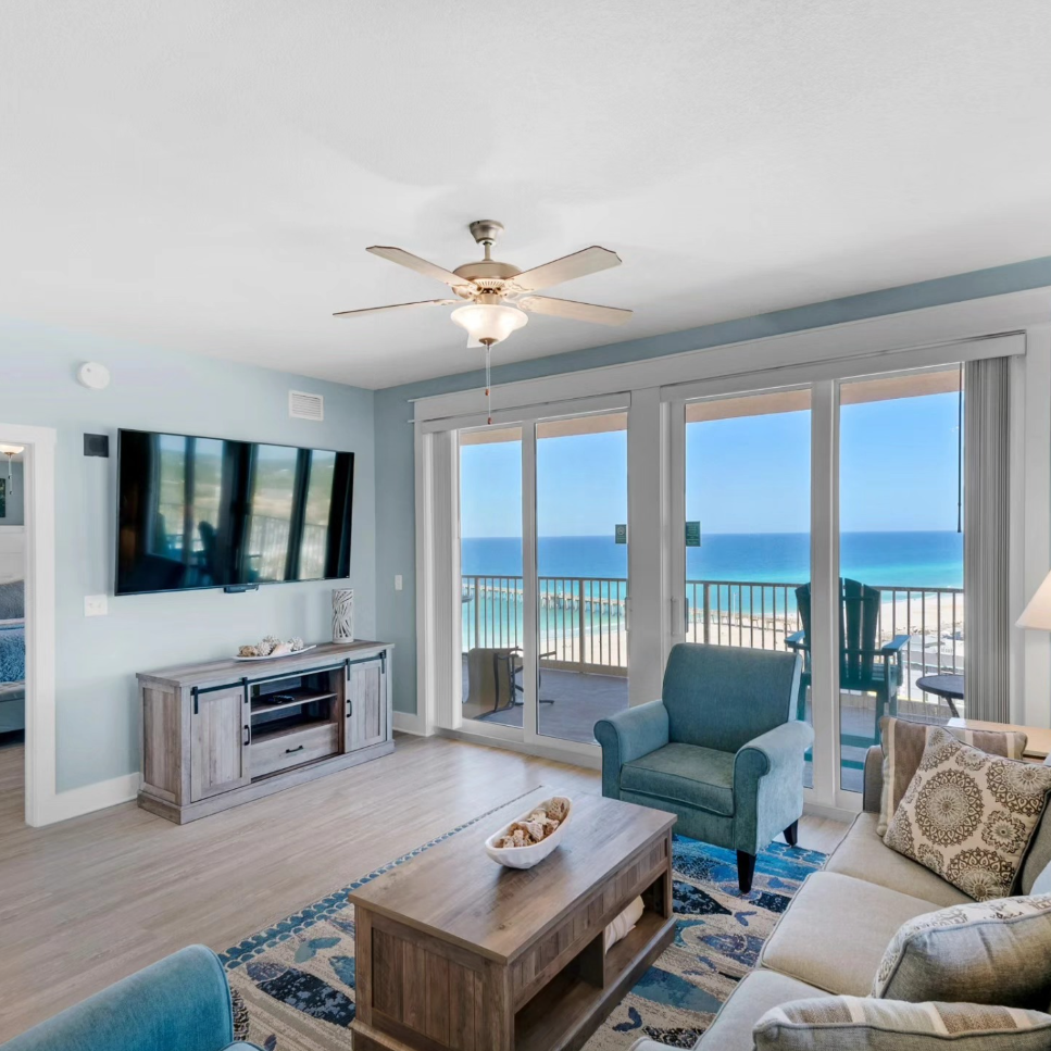A bright, coastal-style living room with a blue armchair, a sofa, and large sliding doors overlooking a sunny ocean view.