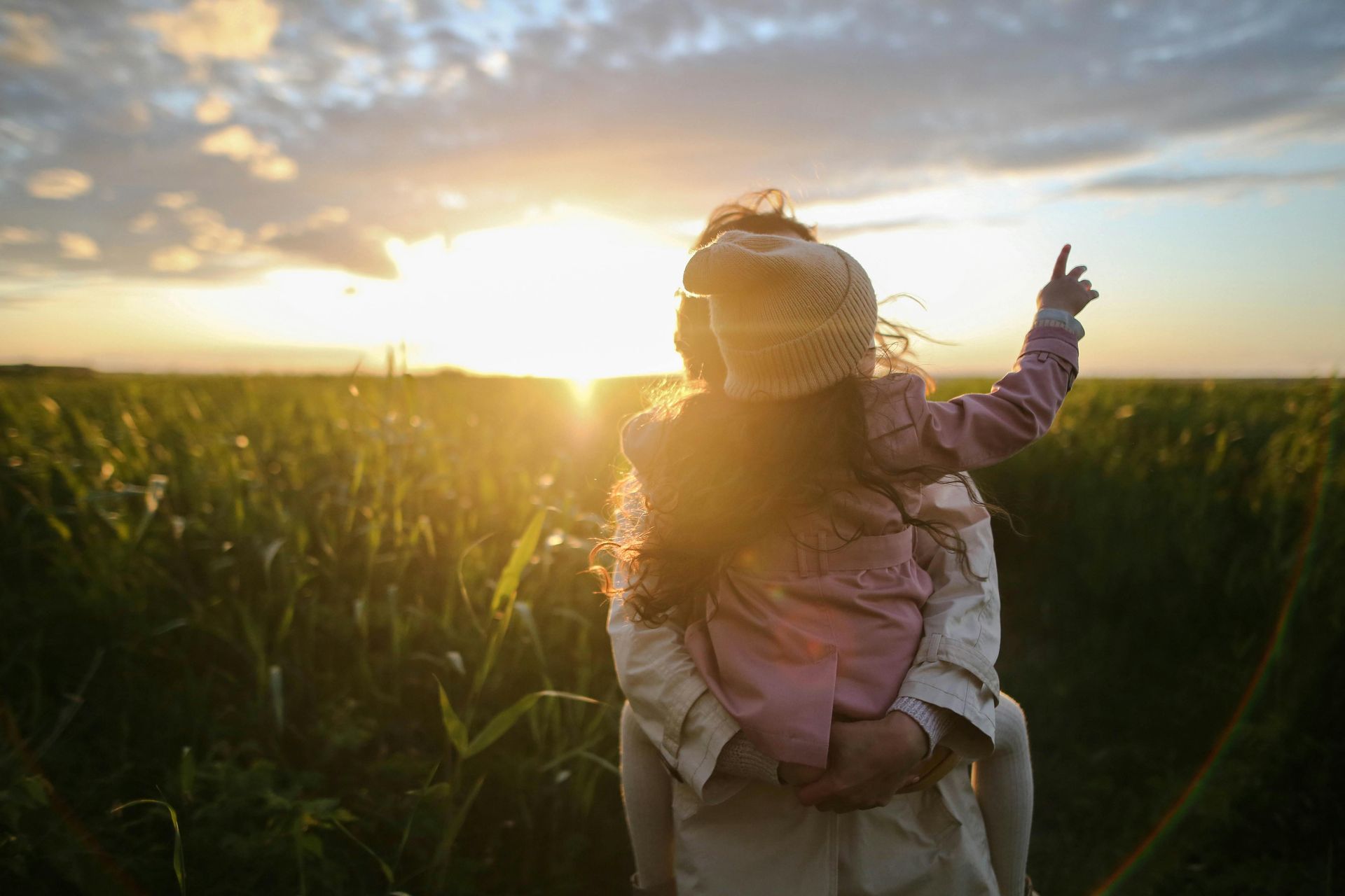 A woman is carrying a little girl on her shoulders in a field at sunset.