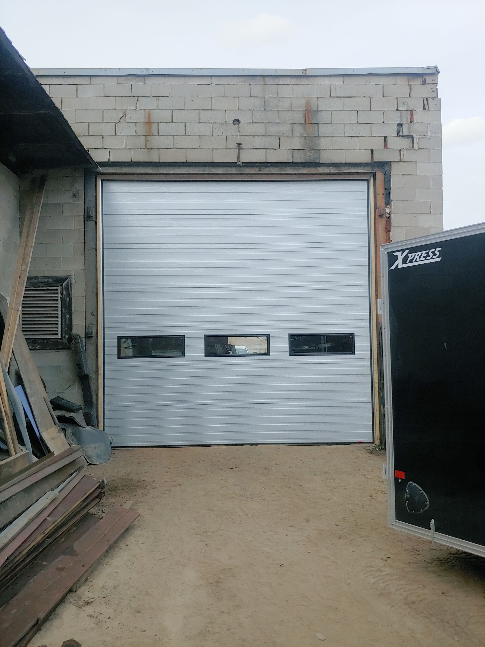 Porte de garage, grise, avec trois petites fenêtres, implantée dans un bâtiment en béton.