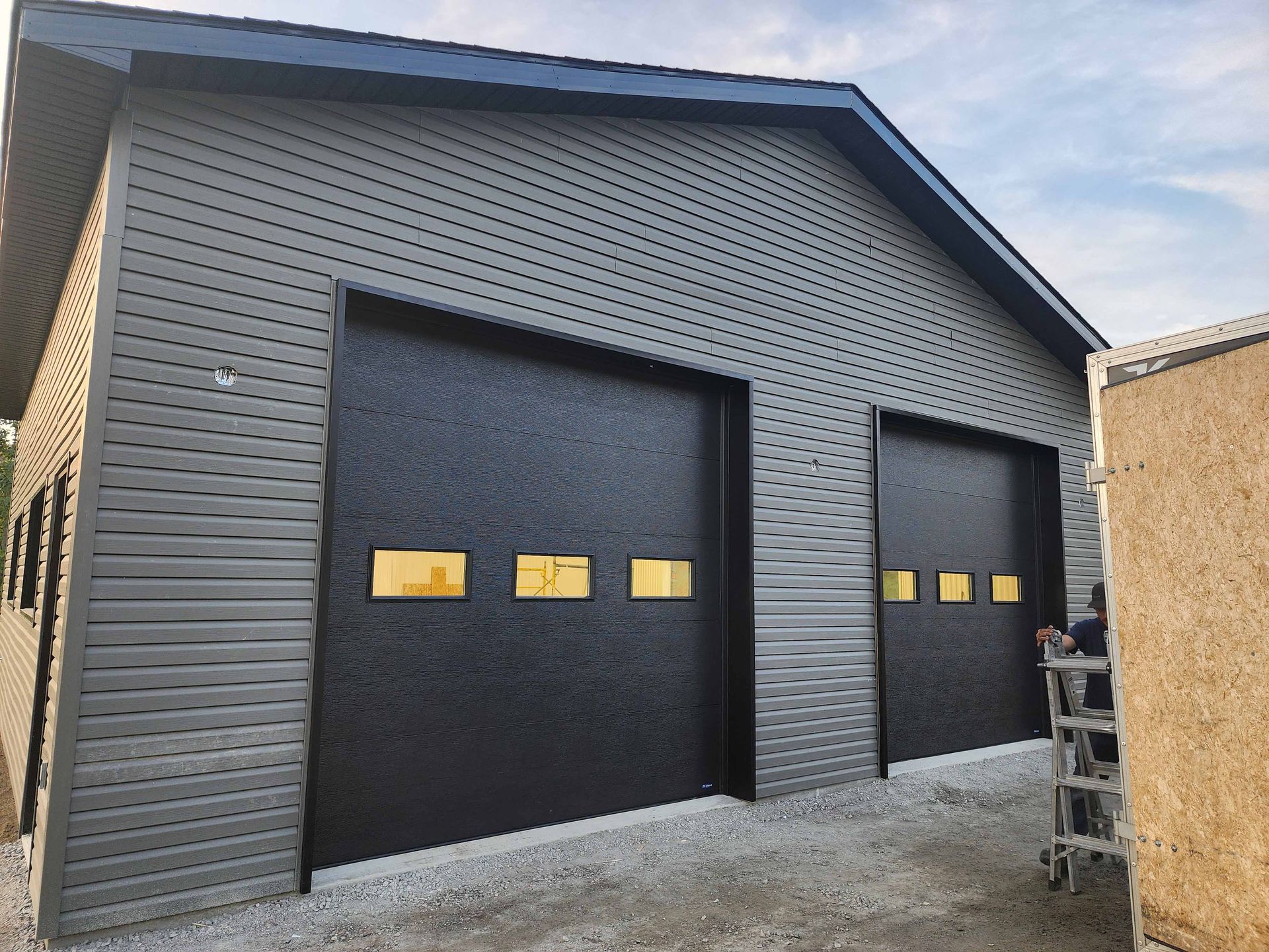 Deux portes de garage noires sur un bâtiment gris avec des fenêtres aux tons dorés.