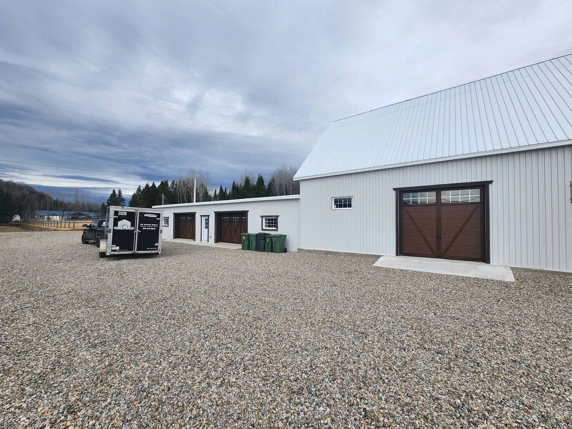 Un bâtiment blanc avec une porte de garage marron se trouve au sommet d'un terrain en gravier.