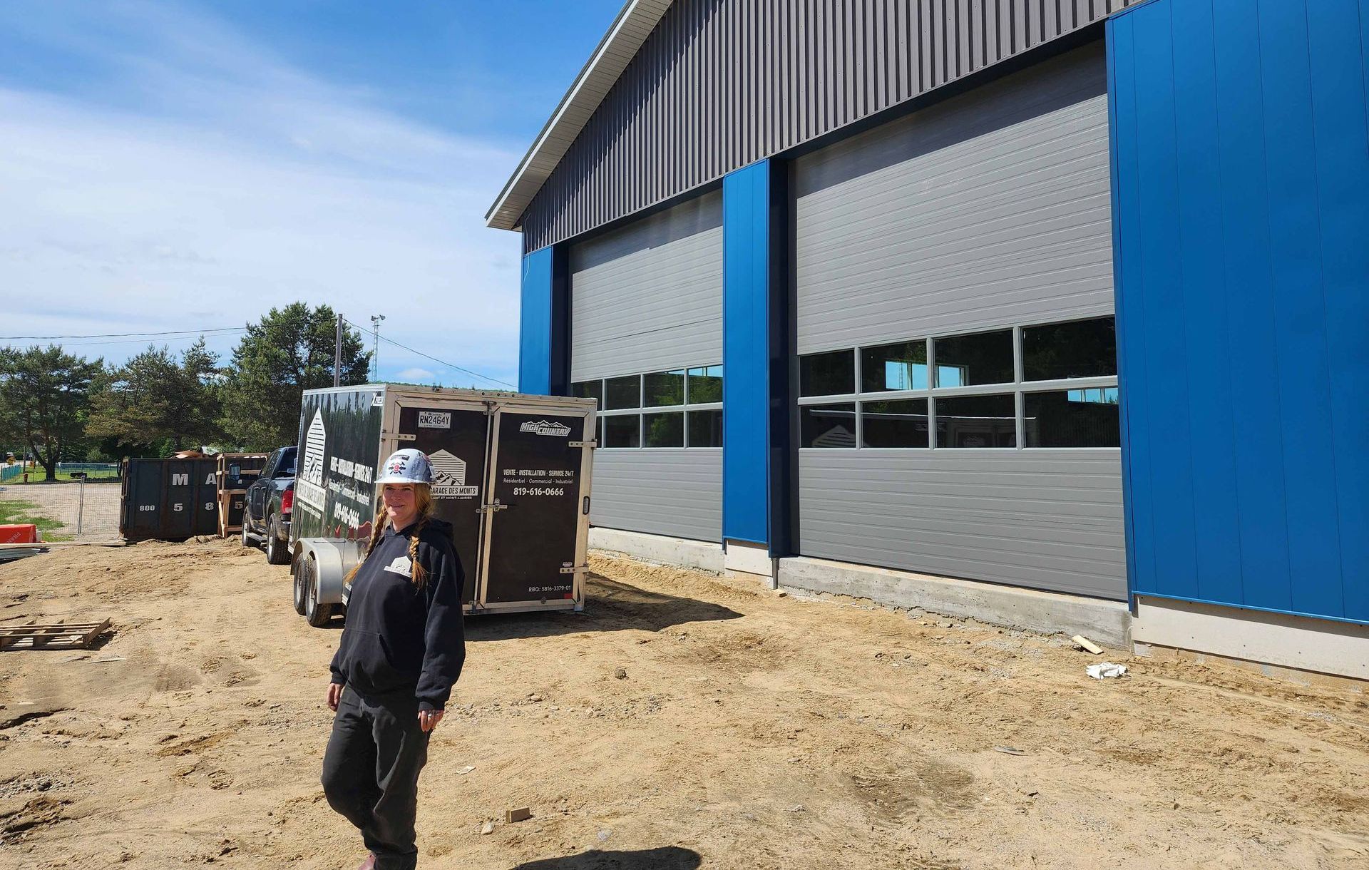Une personne se tient devant un bâtiment  avec de grandes portes de garage et une remorque sur un sol en terre battue.