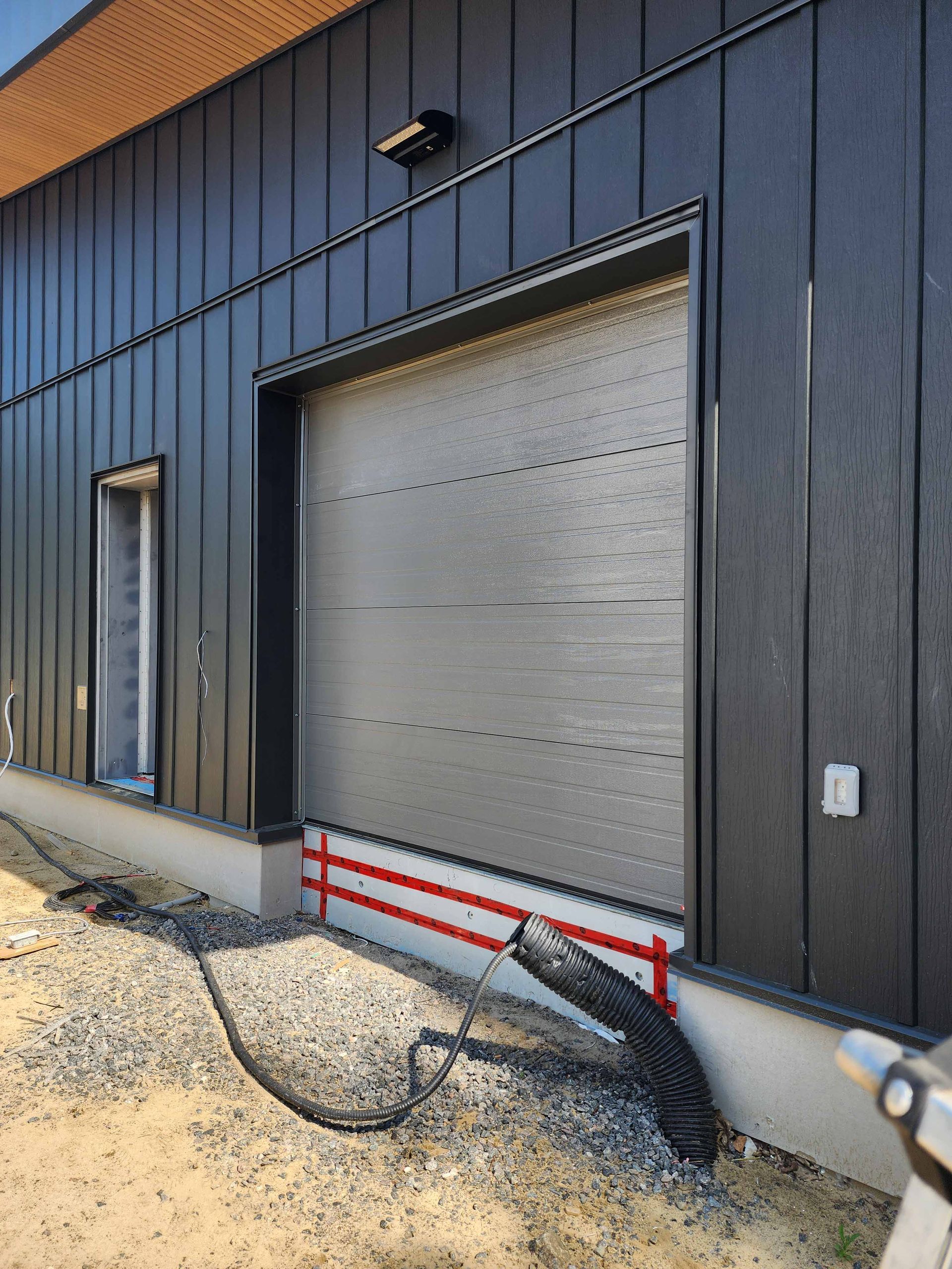 Bâtiment noir avec grande porte en métal, sol en gravier et tuyau exposé.
