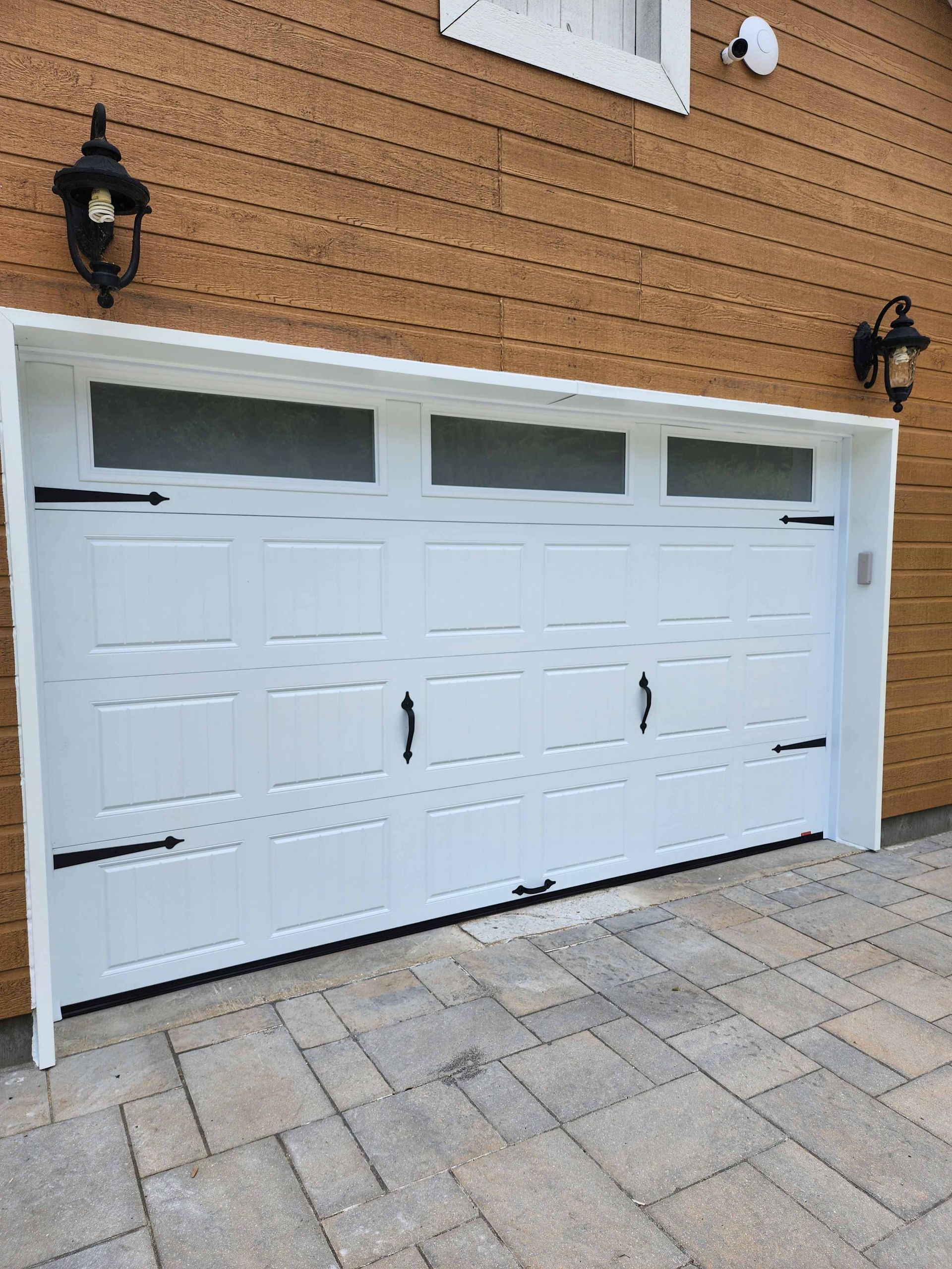 Porte de garage blanche avec quincaillerie noire et trois panneaux en verre dépoli, encastrée dans un mur en bois.