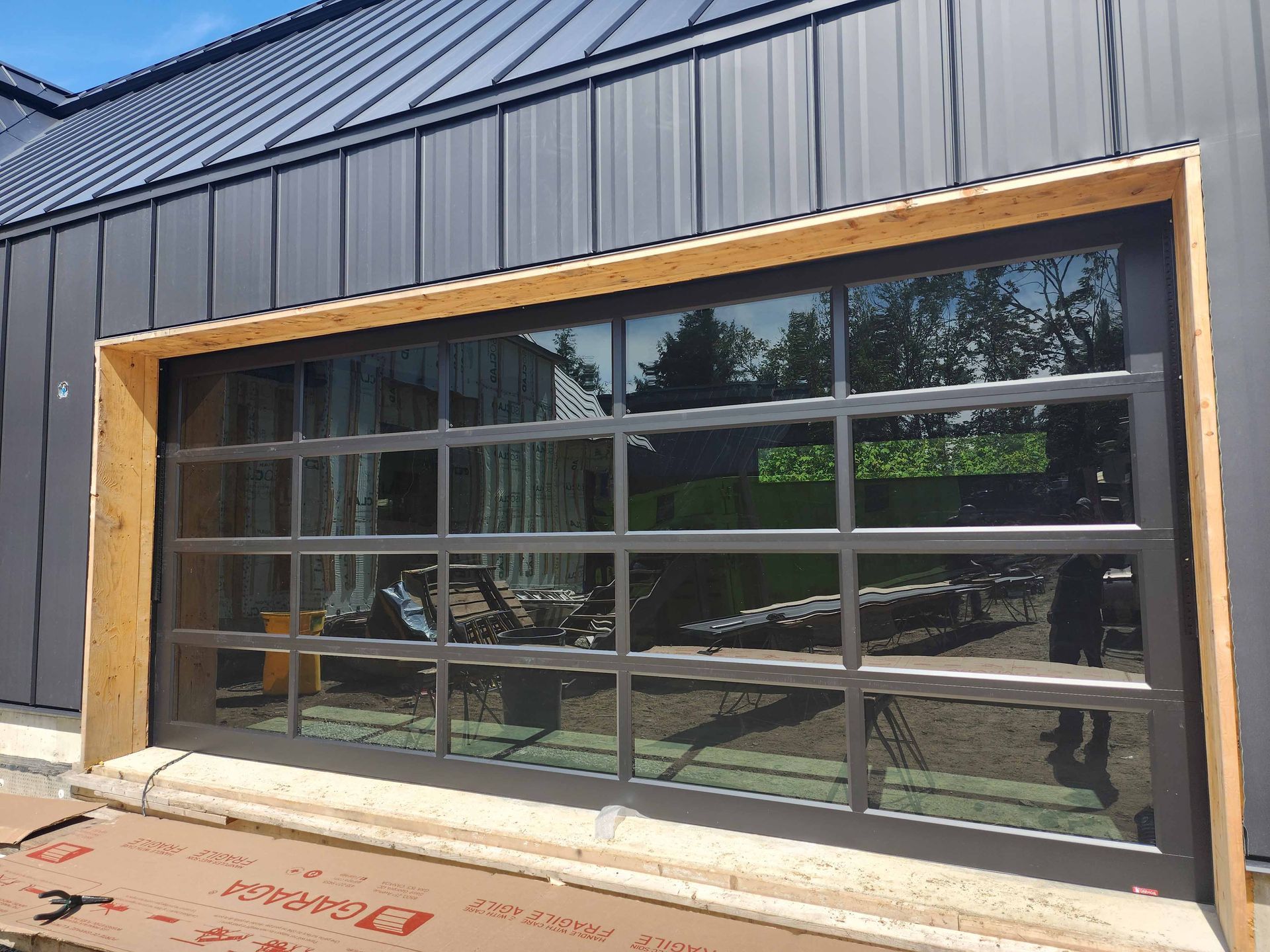 Porte de garage en verre avec cadre gris foncé, se reflétant à l'extérieur, sur fond de bâtiment gris foncé.