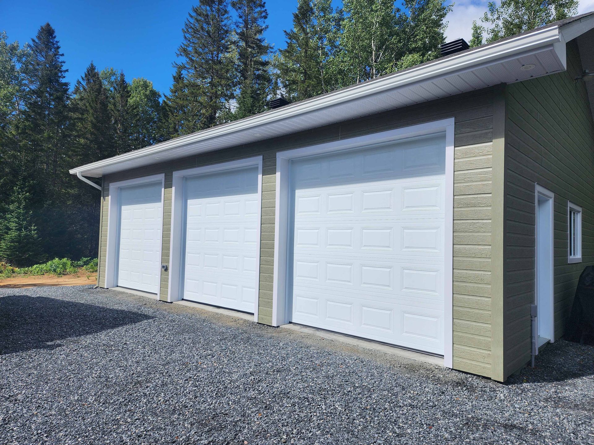 Garage blanc à trois portes avec garniture blanche, gravier gris et arbres en arrière-plan.