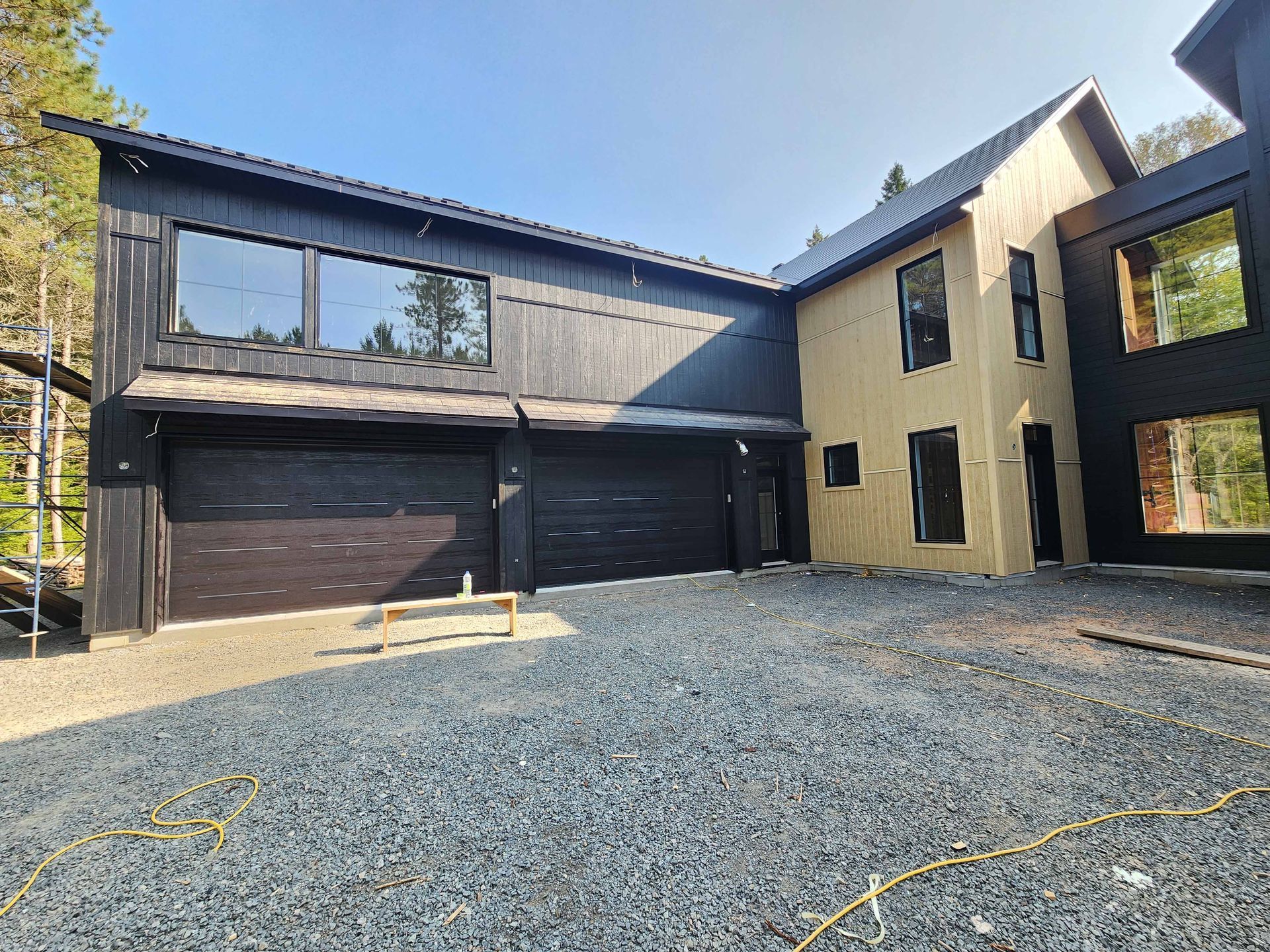 Maison moderne à deux étages avec revêtement noir et beige, garage pour deux voitures, allée en gravier et grandes fenêtres.