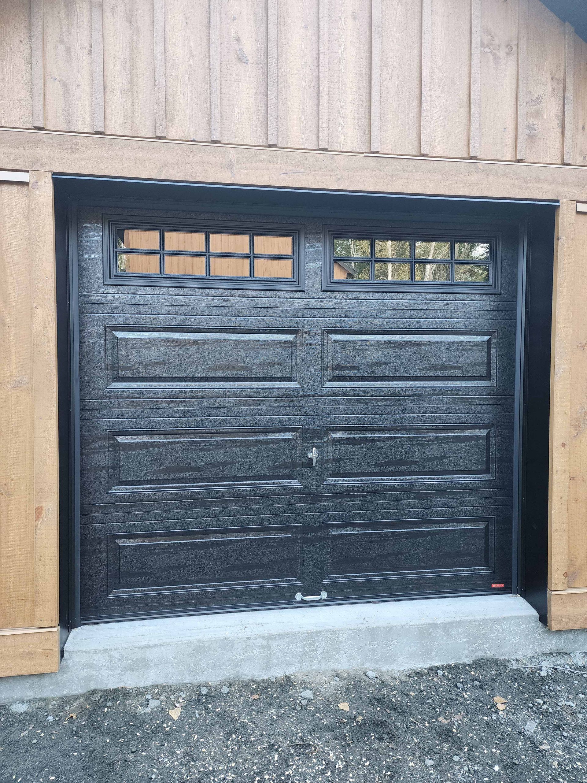 Une porte de garage noire avec une fenêtre se trouve devant un bâtiment en bois.