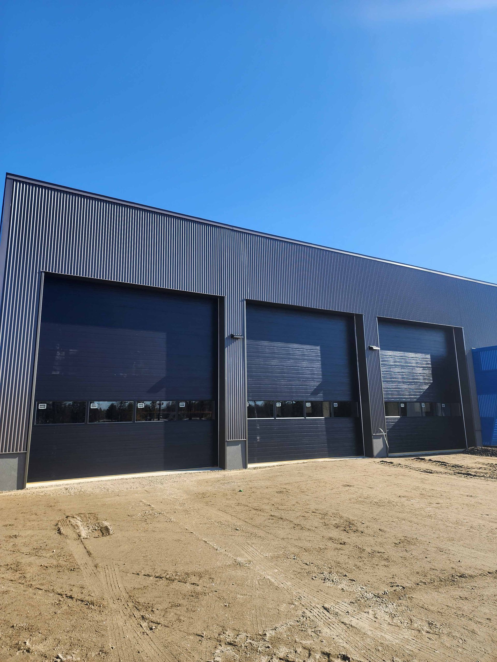 Trois portes de garage noires sur un bâtiment avec revêtement en tôle ondulée, par une journée ensoleillée.