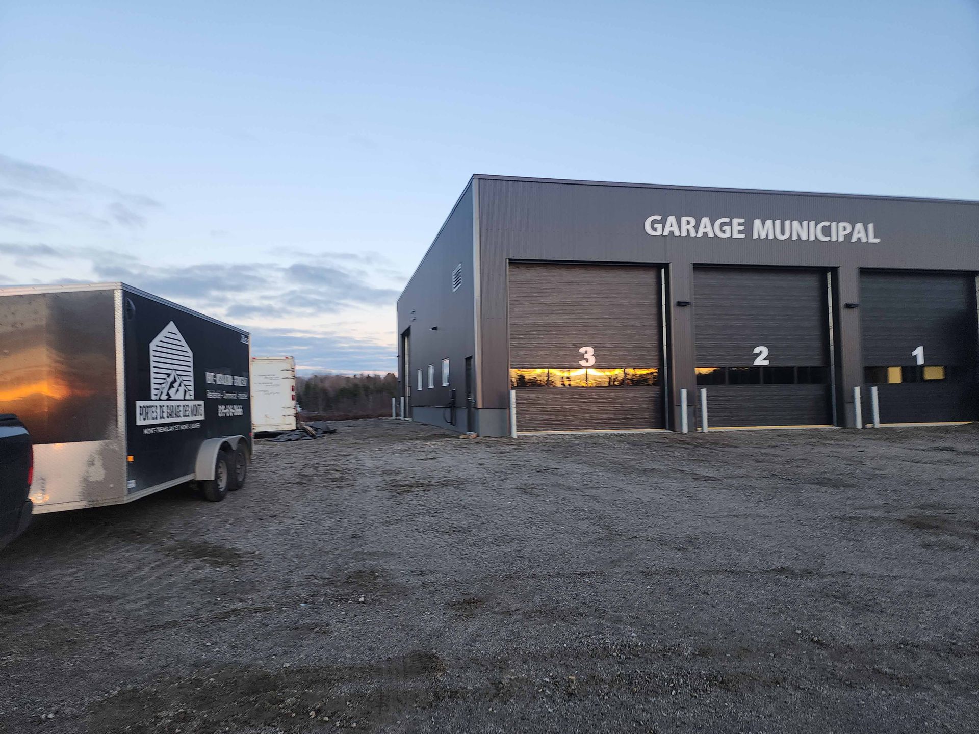 Vue extérieure d'un garage municipal avec des emplacements numérotés, une remorque et un parking en gravier.