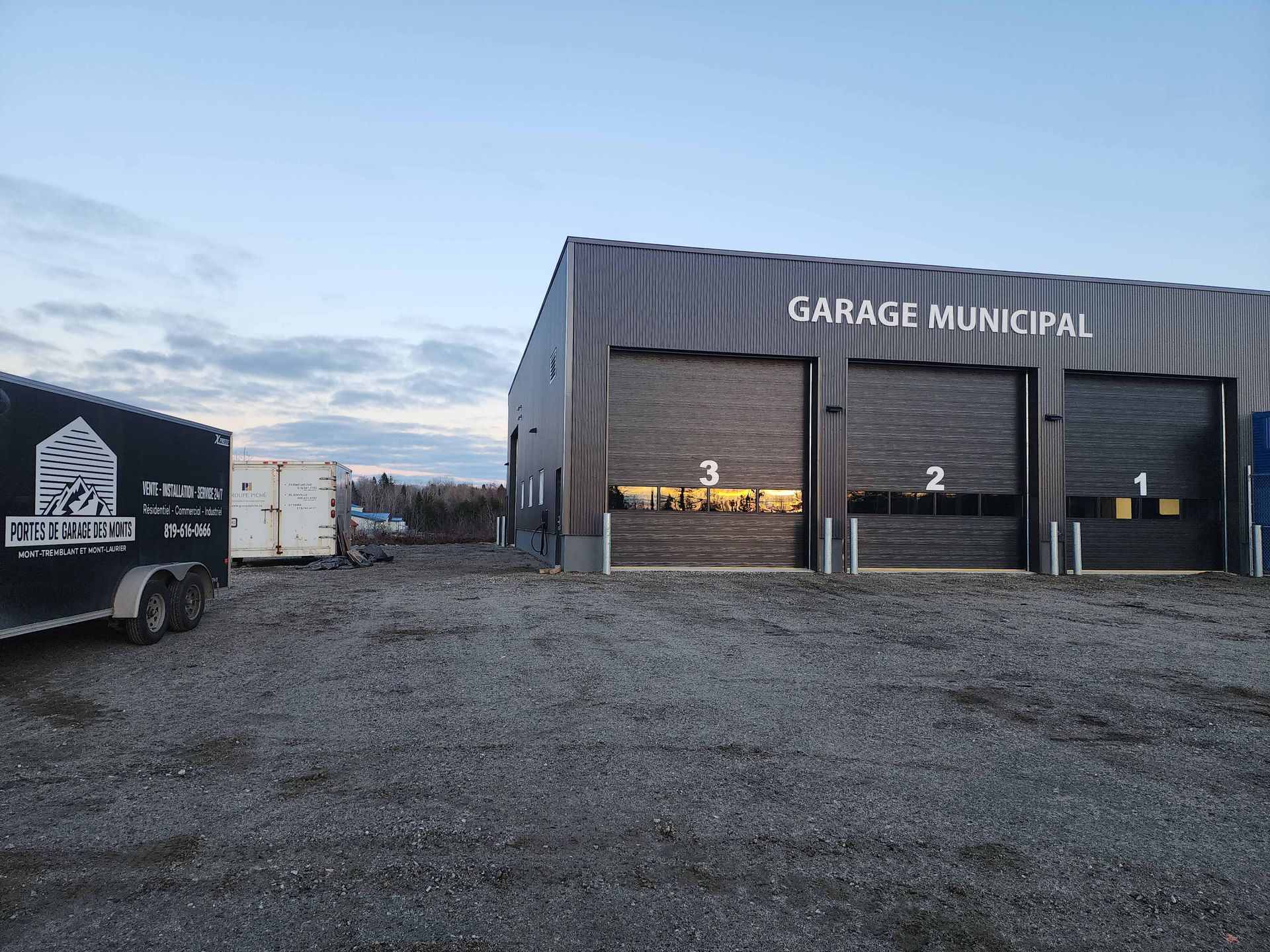 Garage municipal avec trois portes numérotées, façade foncée, terrain en gravier, ciel bleu.