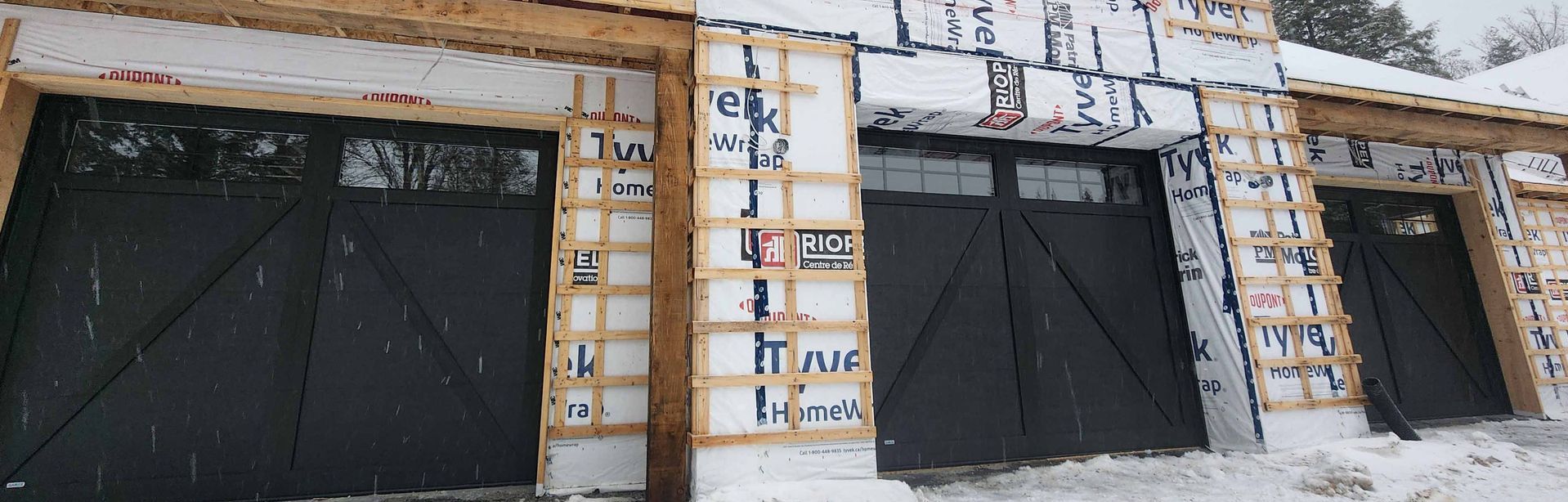 Vue intérieure d'un garage en construction, avec trois portes noires fermées. On aperçoit les câbles électriques jaunes.