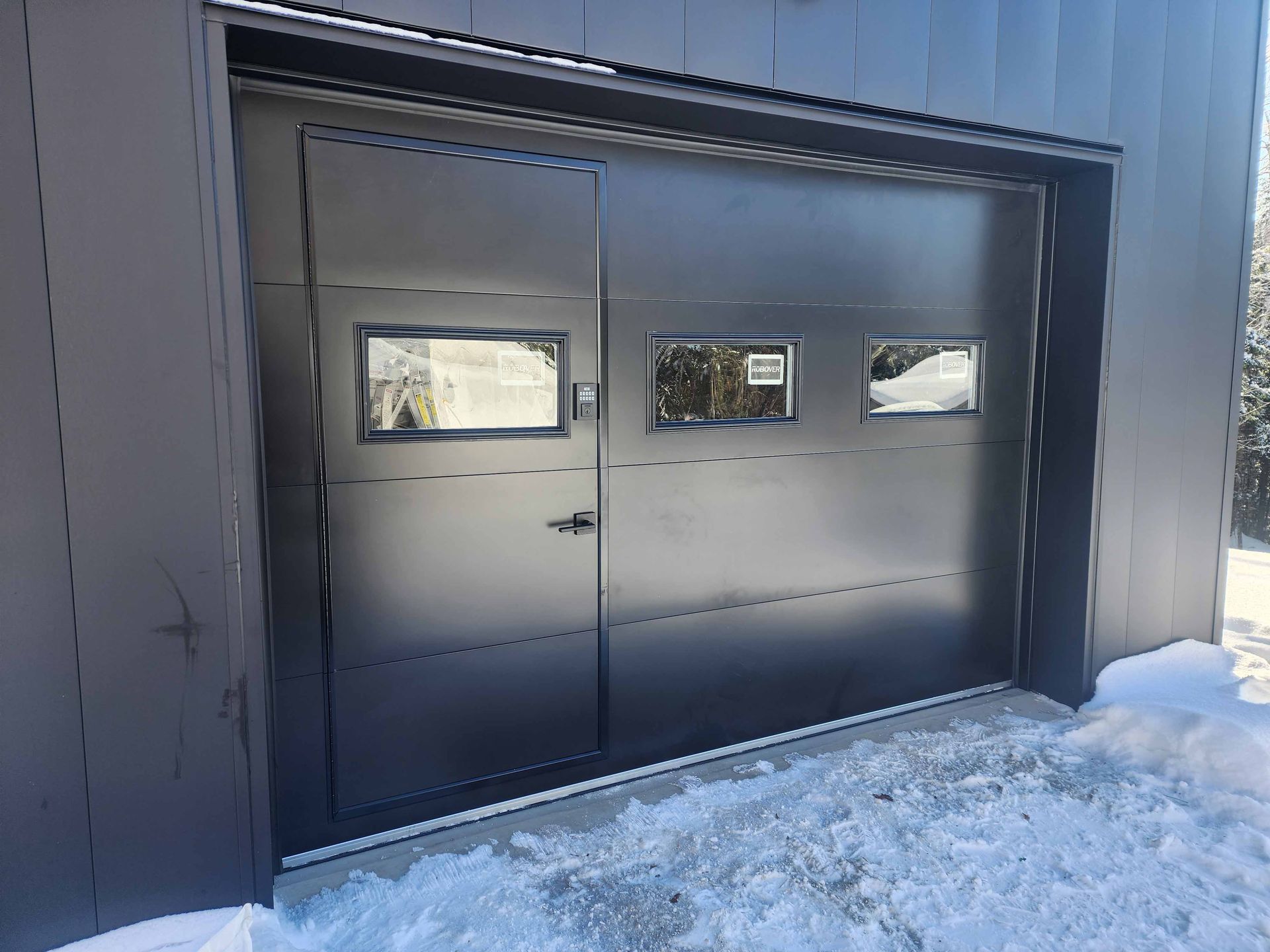 Porte de garage grise avec trois fenêtres, neige au sol.