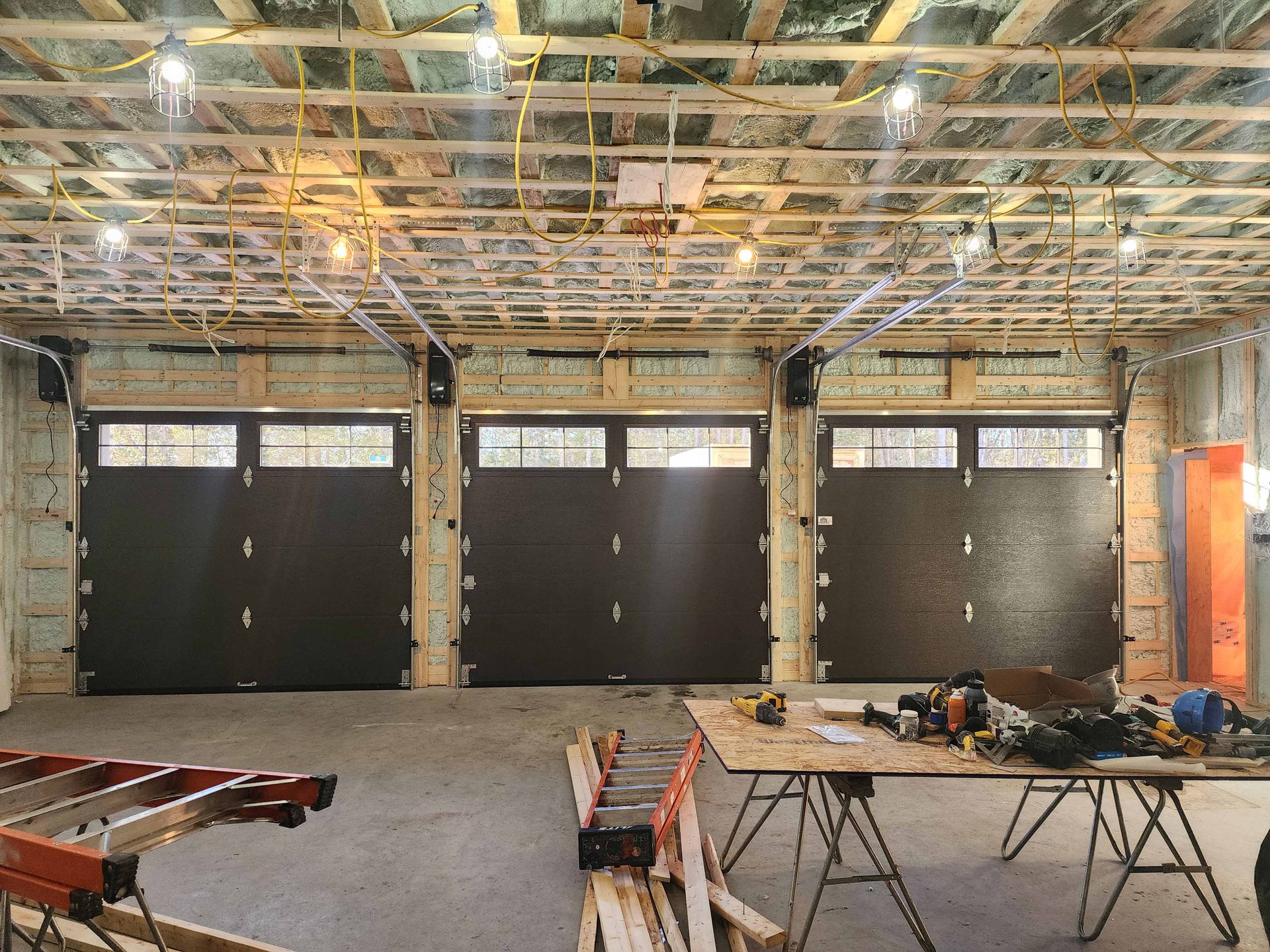 Vue intérieure d'un garage en construction, avec trois portes noires fermées. On aperçoit les câbles électriques jaunes.