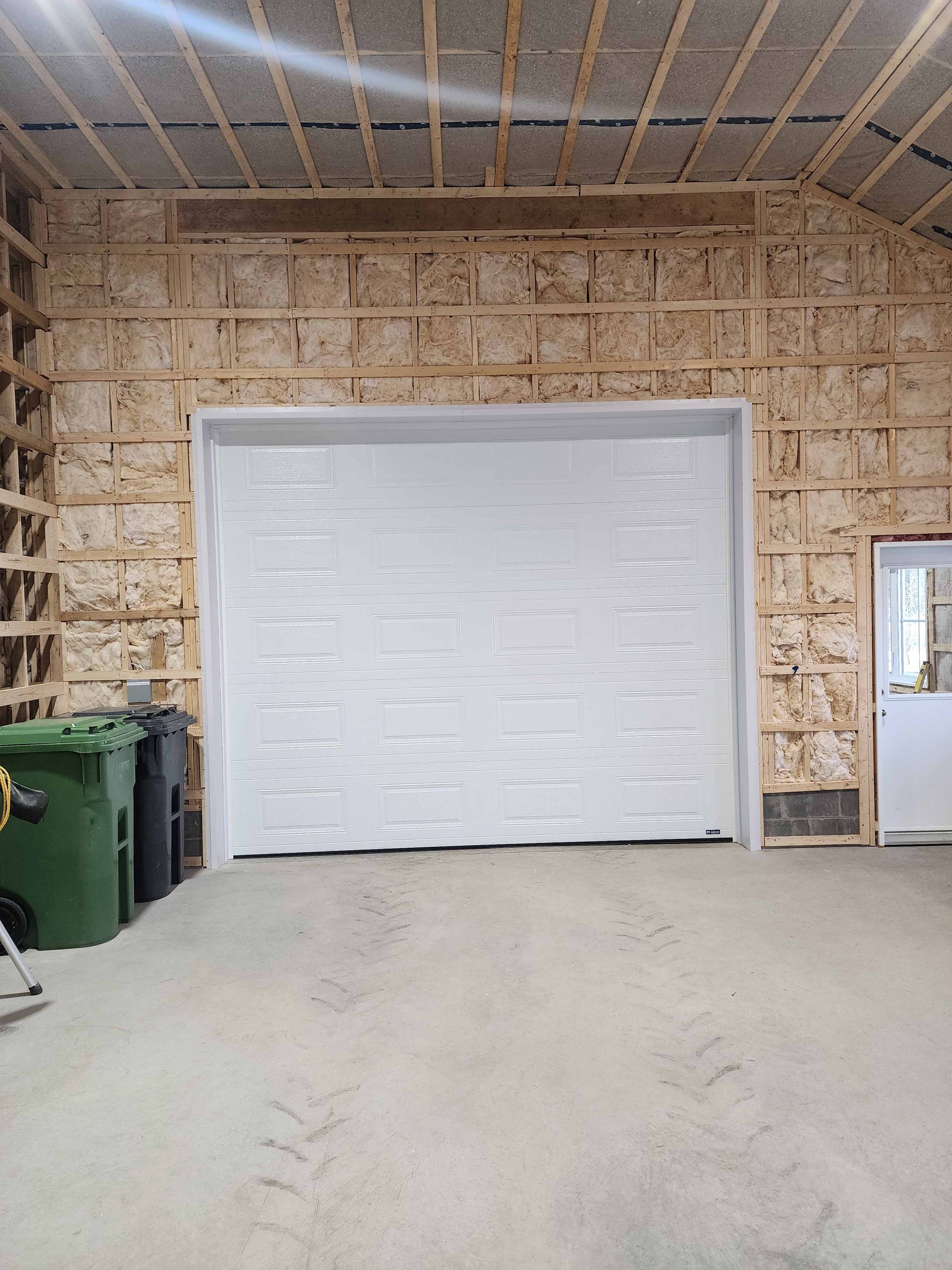 Un garage avec une porte de garage blanche et une poubelle verte.