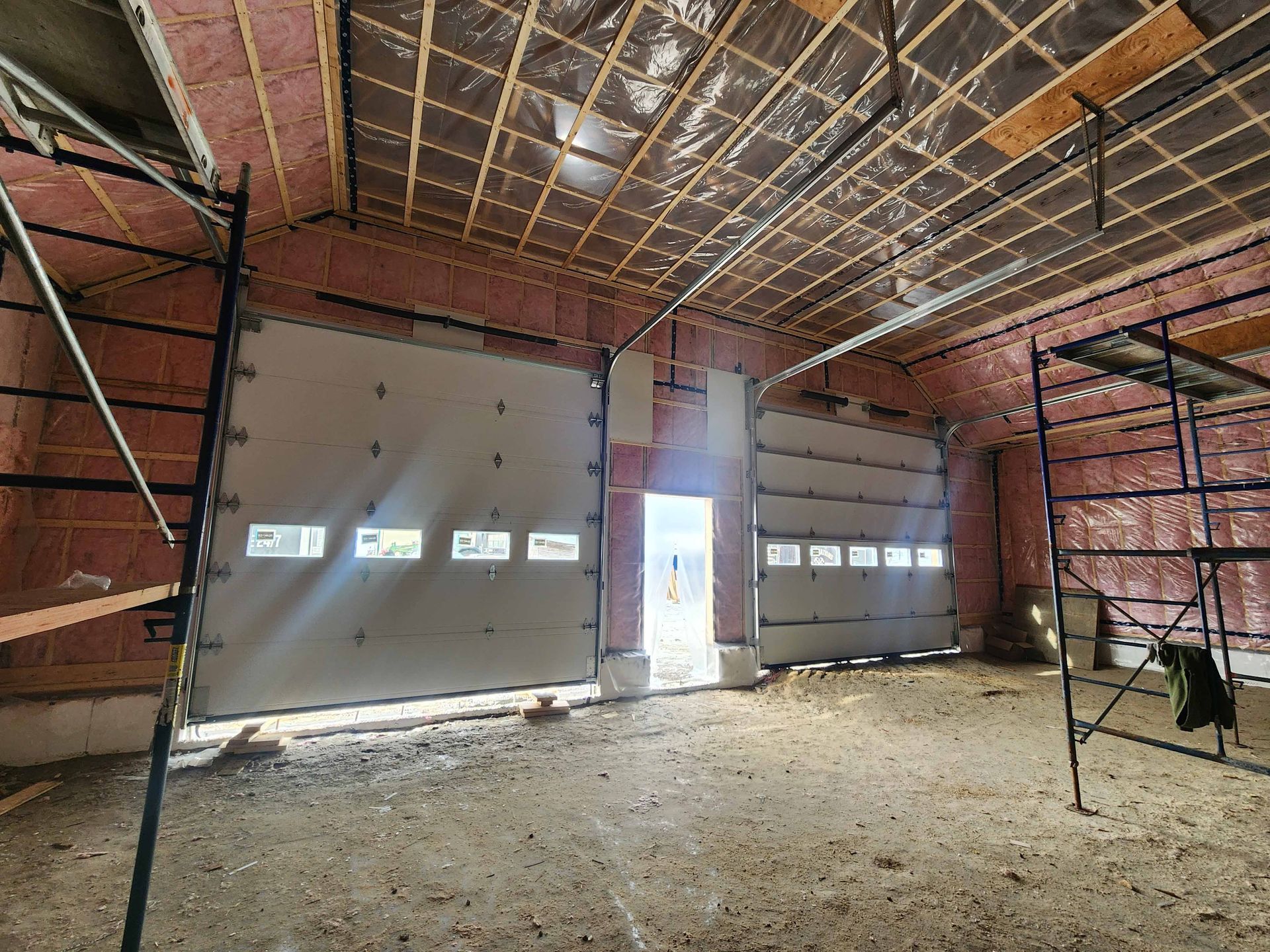 Intérieur d'un bâtiment en construction, montrant des portes de garage, de l'isolant et des poutres de plafond apparentes.