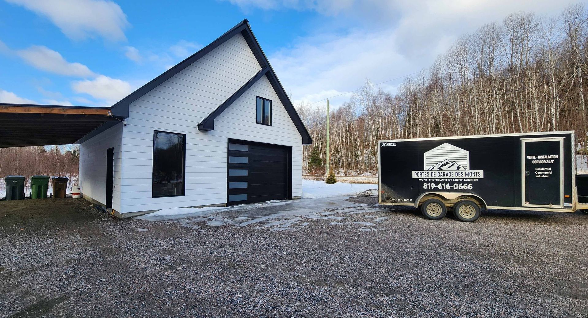 Bâtiment blanc avec une porte noire à côté d'une remorque noire sur une allée de gravier, sous un ciel bleu.