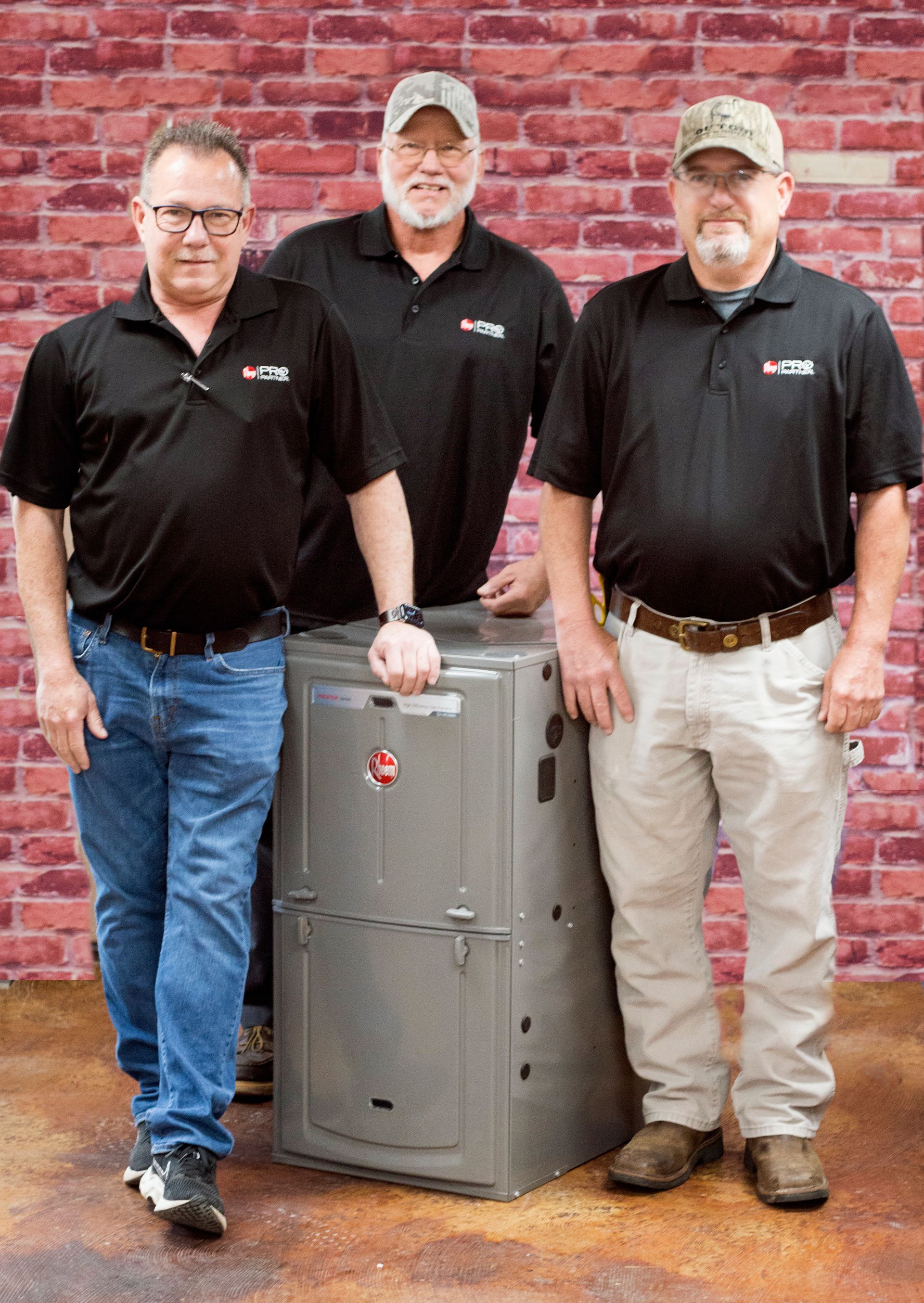 Three HVAC technicians pose with a furnace against a brick wall. They wear black shirts and look at the camera.