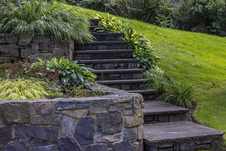 Natural stone retaining wall and stairway surrounded by lush green landscaping, ornamental grasses, and garden plants on a sloped backyard hillside.