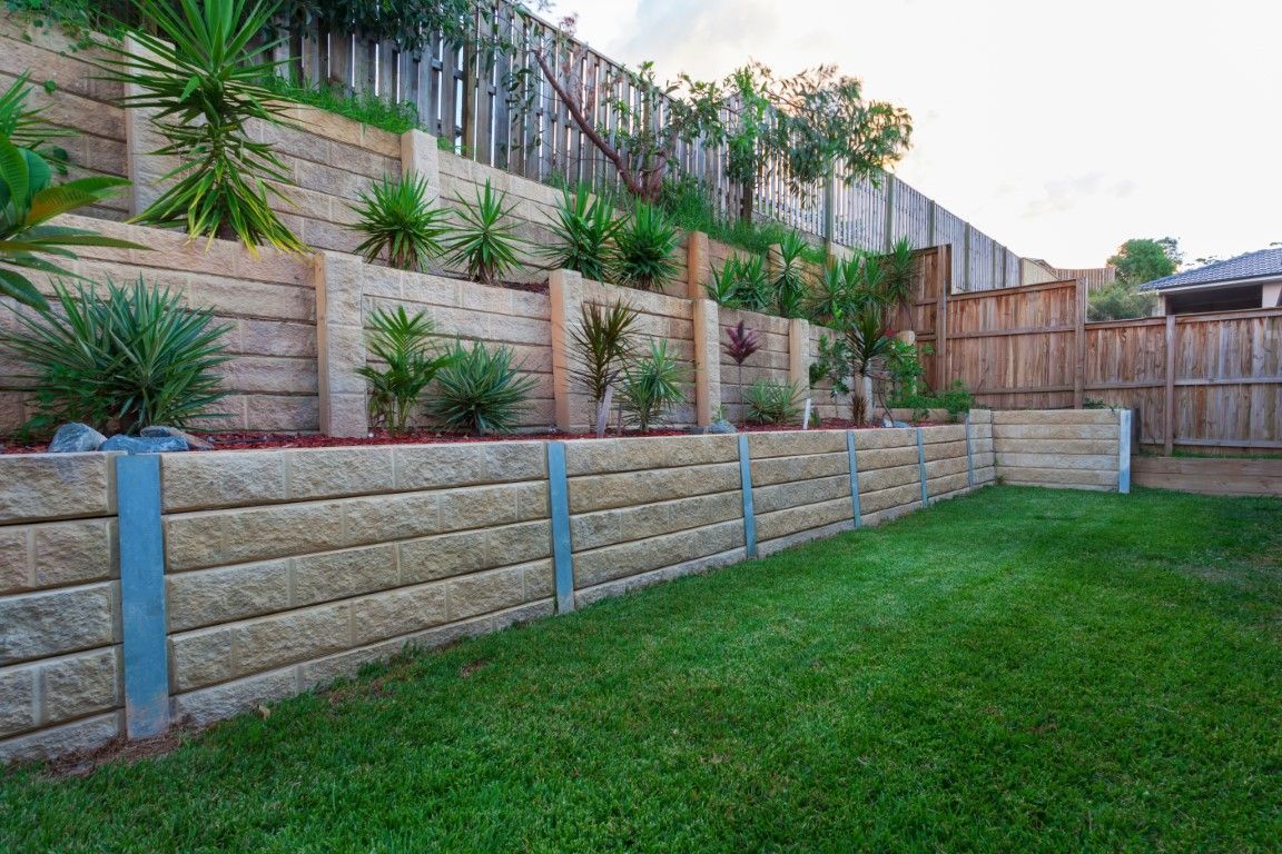 Retaining walls of stacked beige blocks support tiered garden beds with green plants, next to a grass lawn and wooden fence.