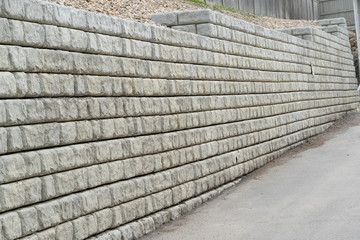 Retaining wall made of gray concrete blocks. The wall slopes upward and is located next to a road.