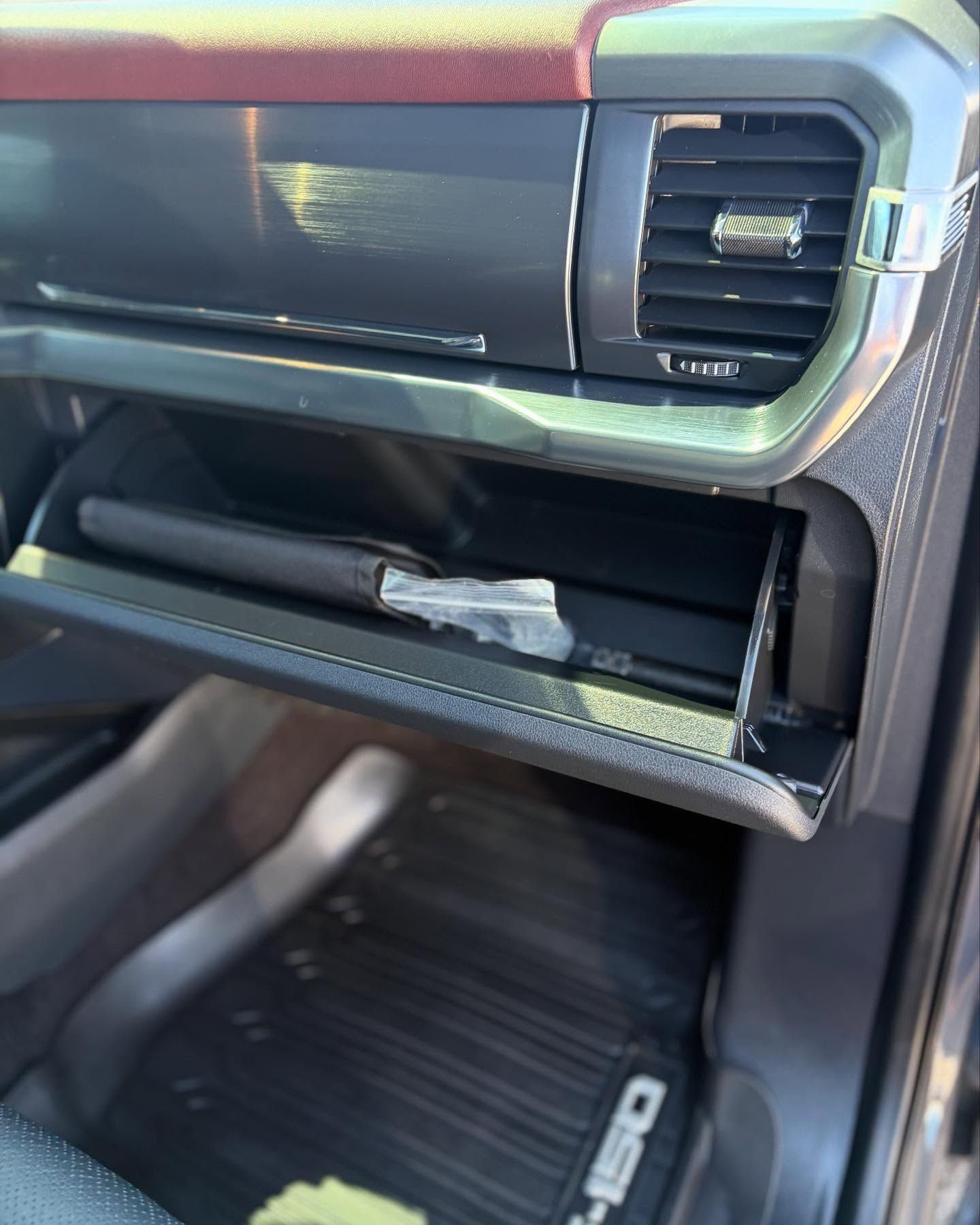 Open glove box of a Ford F-150 truck with a dark interior, featuring a red trim on the dashboard and a rubber floor mat.