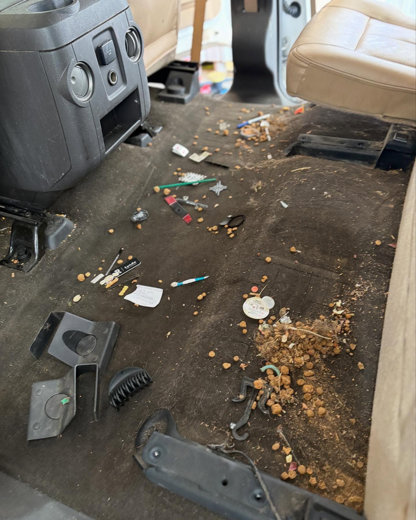 A messy, dark car floor covered in loose dirt, debris, cigarette butts, and plastic pieces between two tan car seats.