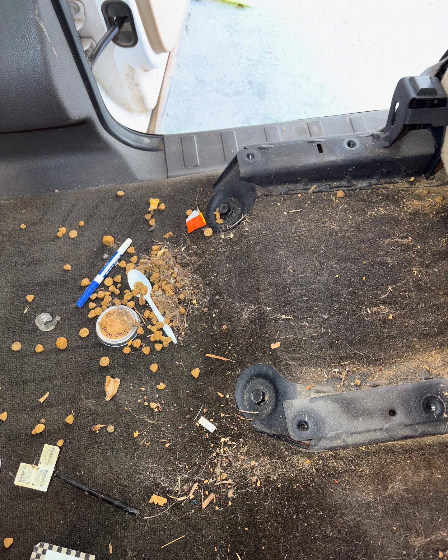 A dirty vehicle interior floor with spilled food, a plastic spoon, a blue pen, and debris scattered near the seat brackets.