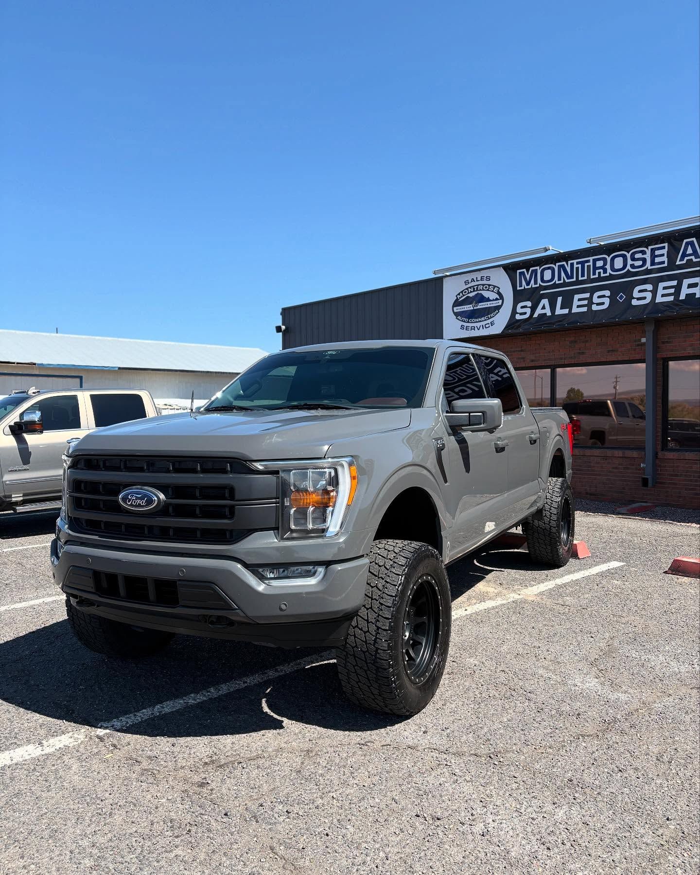 A gray Ford F-150 truck with a lifted suspension and off-road tires parked in front of an automotive sales building.