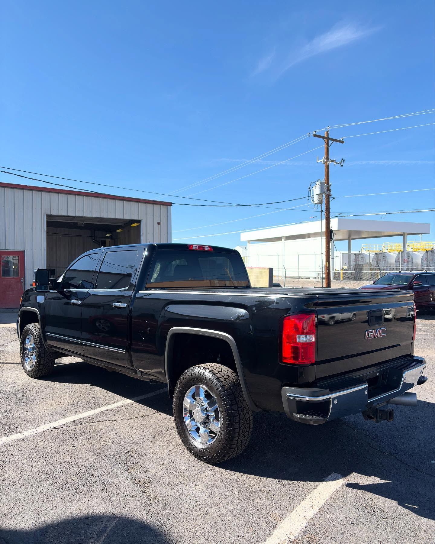 A black GMC pickup truck parked in a gravel lot on a sunny day with a service building and power pole in the background.