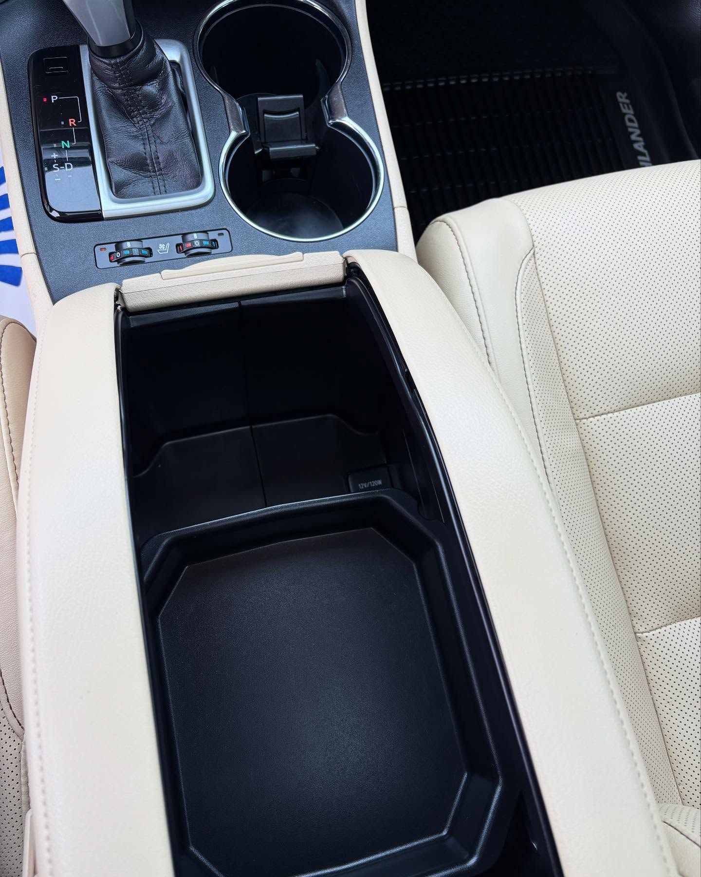 Open center console compartment in a vehicle with beige leather interior, cup holders, and a gear shift.