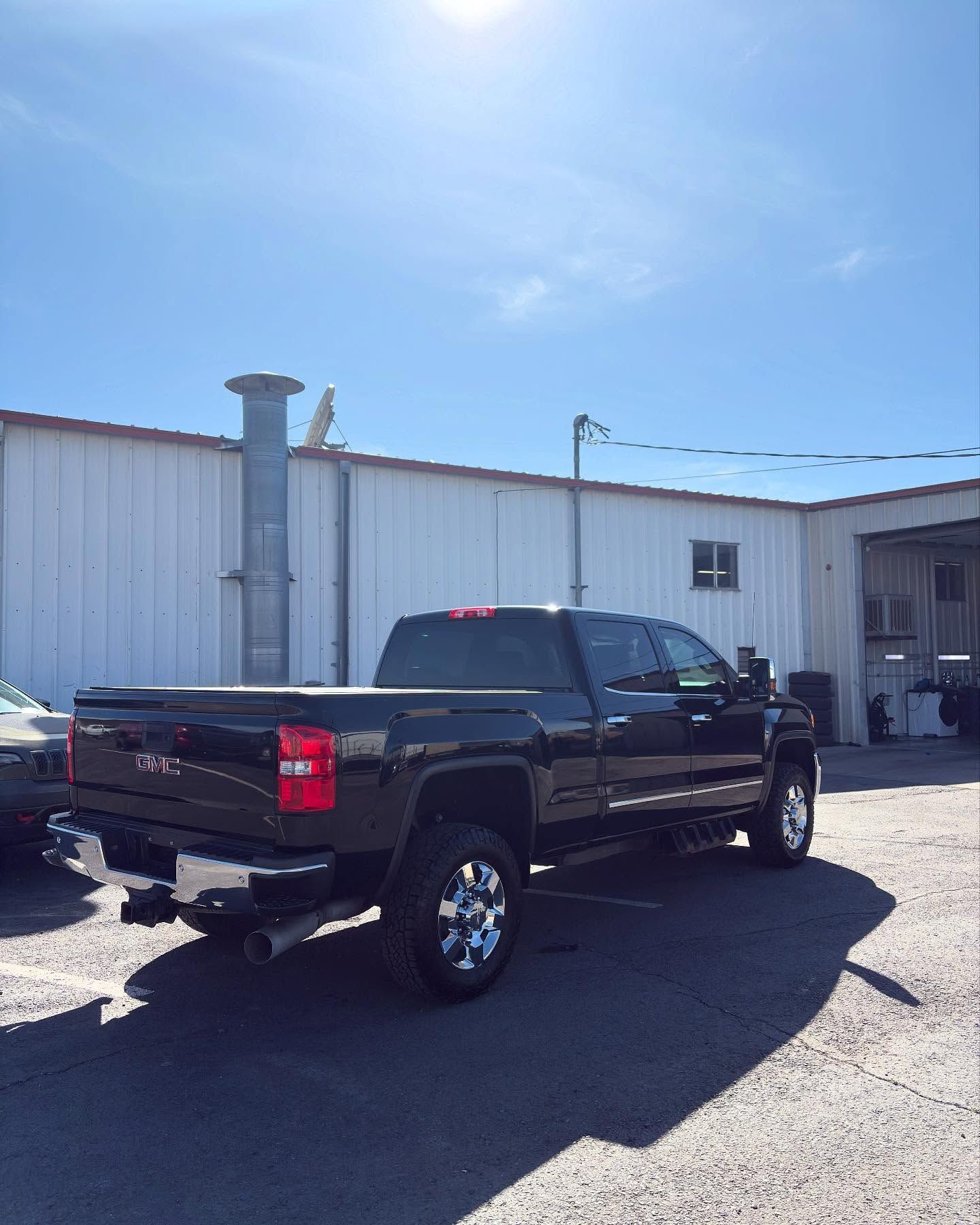 A black GMC pickup truck parked in a gravel lot next to a white metal building under a bright blue sky.