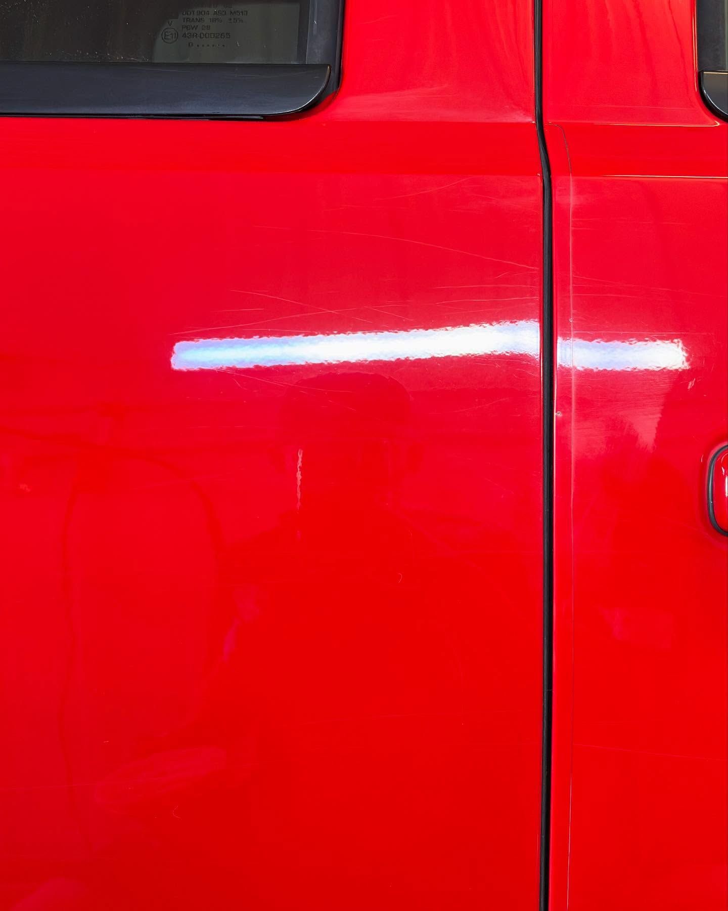 A close-up of a bright red car door with a vertical seam and a light reflection visible on the paint surface.
