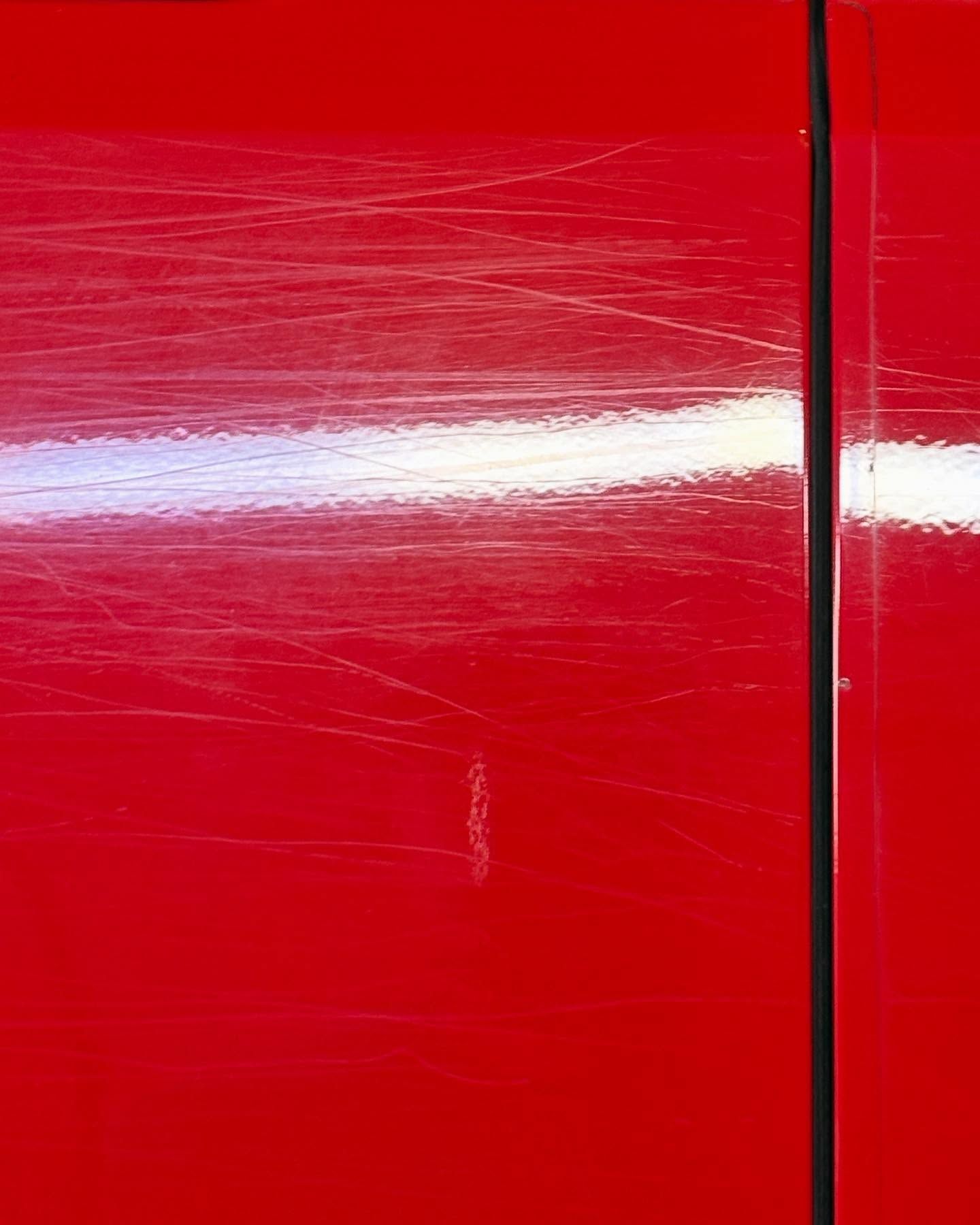 A close-up of a bright red car panel featuring numerous fine scratches and swirl marks across the paint surface.