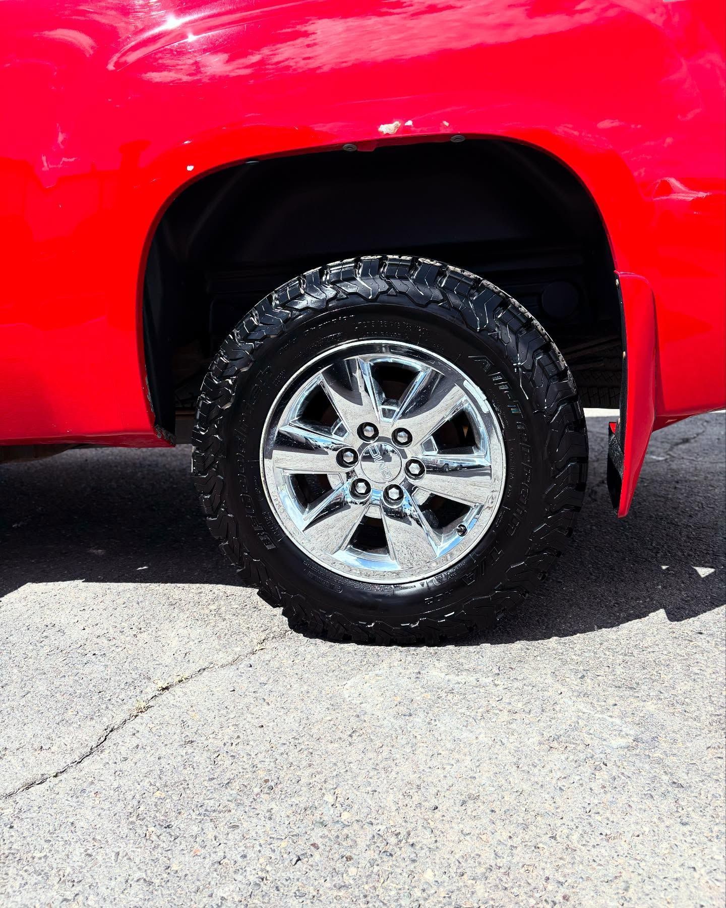 A close-up view of a bright red truck's rear wheel, featuring a chrome rim with an all-terrain tire on asphalt.