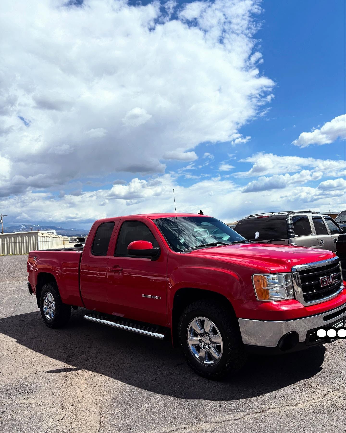 A bright red GMC pickup truck parked on a gravel lot under a blue sky with fluffy white clouds.