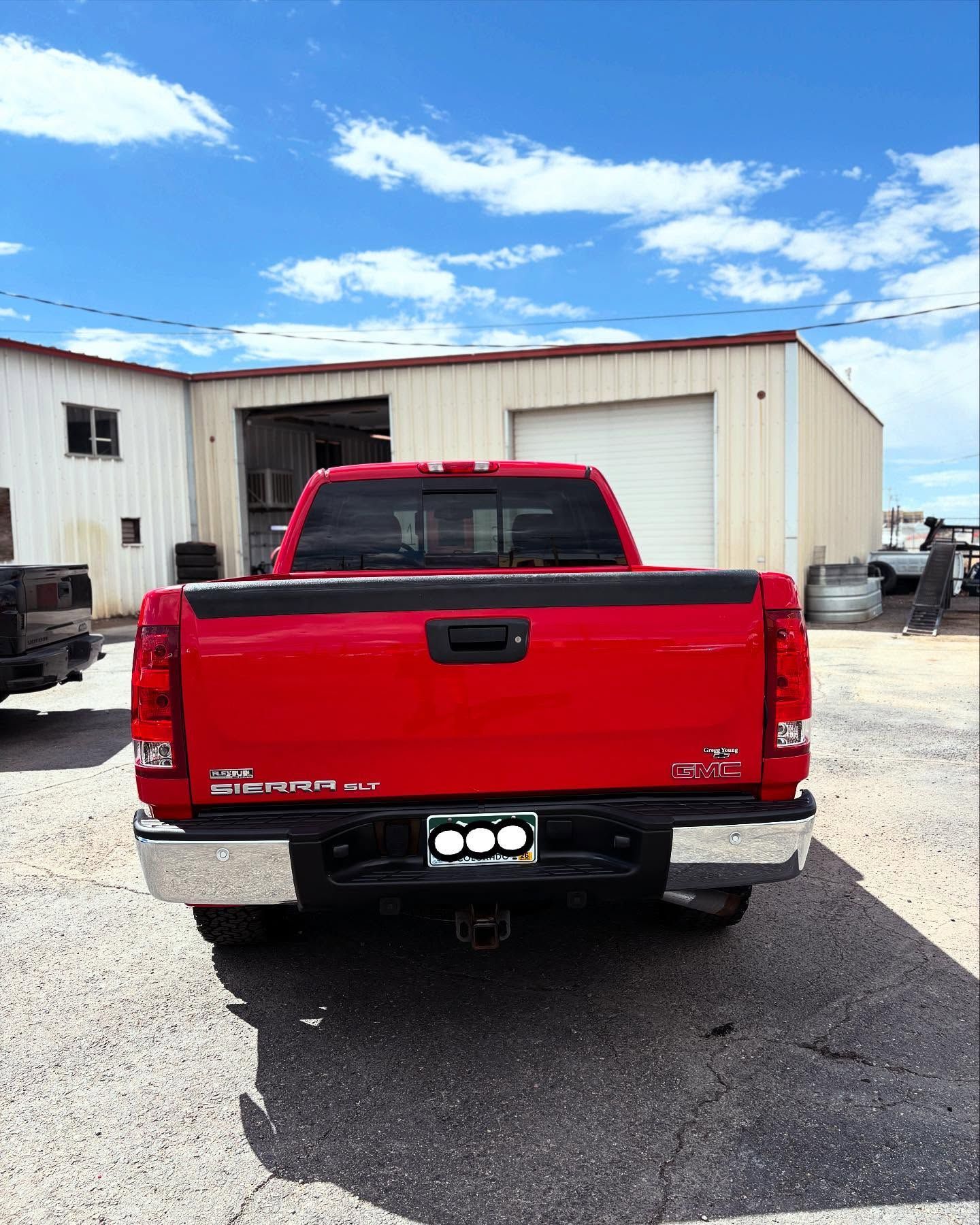 Rear view of a red pickup truck parked on a paved lot in front of a white metal building under a blue, cloudy sky.