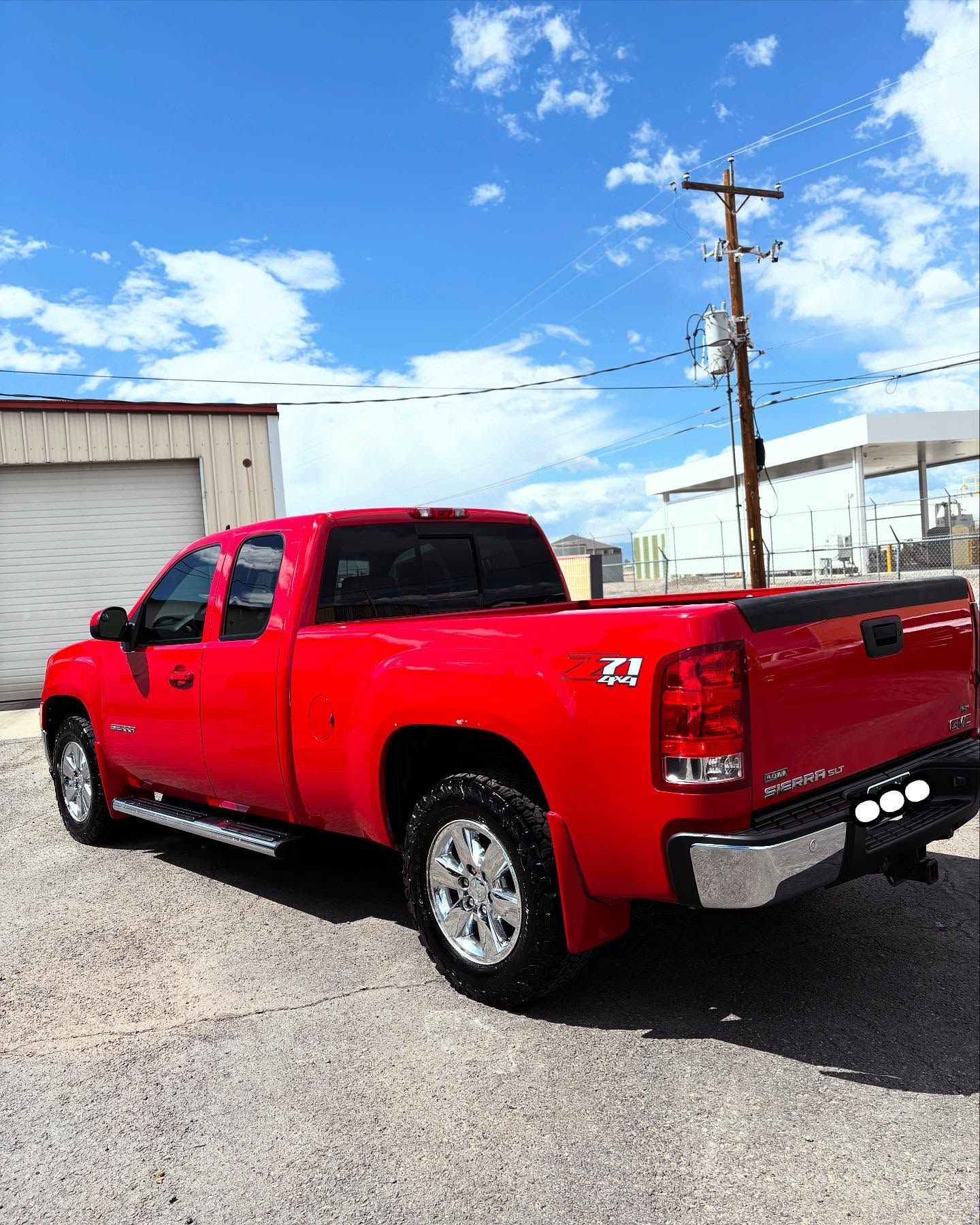 A red extended cab pickup truck parked on a gravel lot under a bright blue sky with clouds.