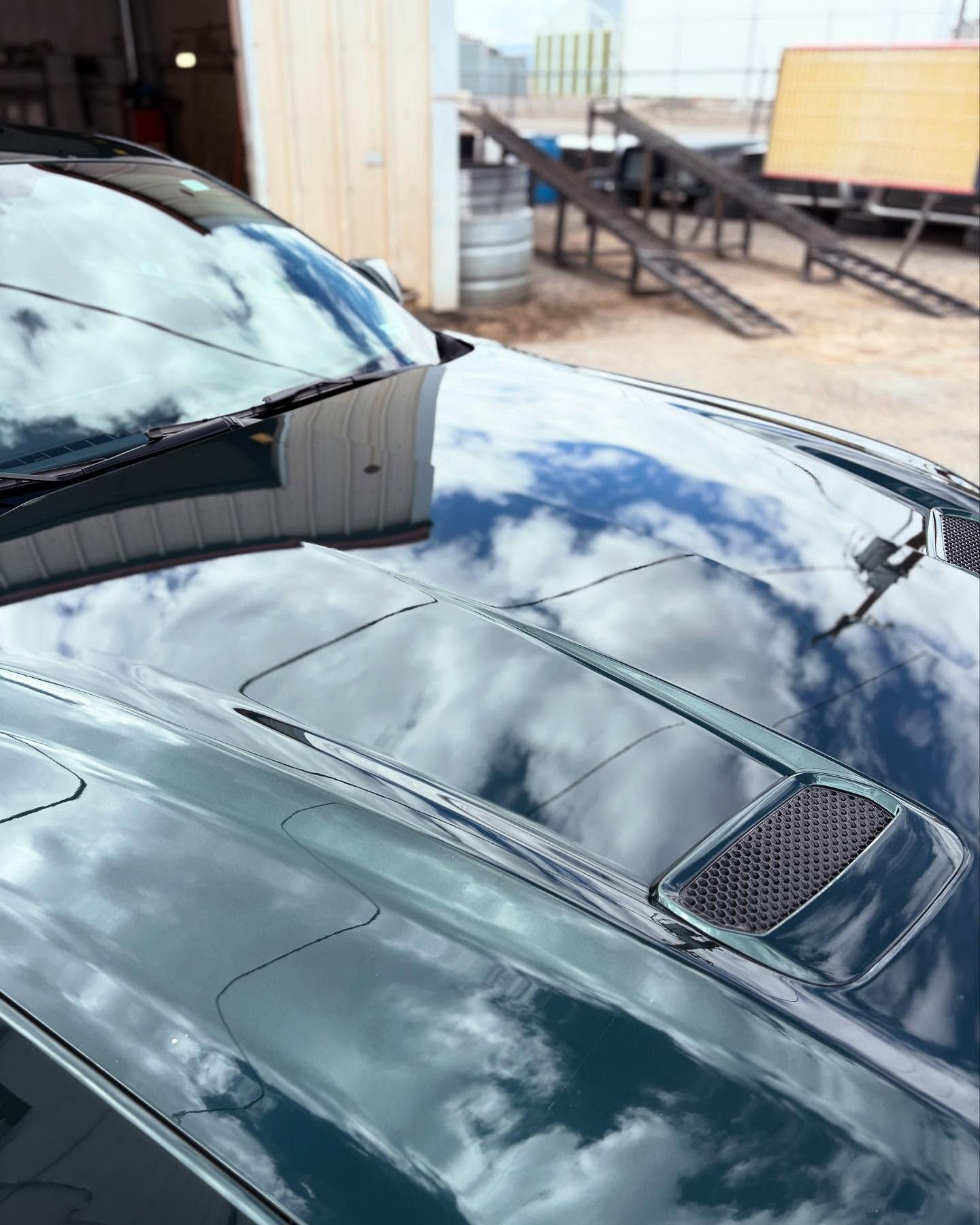 High-angle shot of a dark green car hood with a black mesh vent and sky reflections, parked in a garage area.