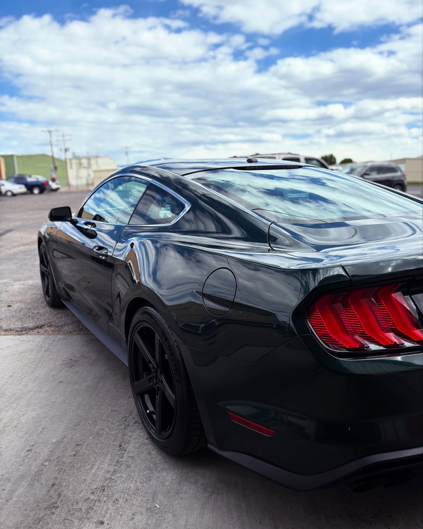 A black Ford Mustang parked in an outdoor lot under a cloudy blue sky, viewed from the rear passenger side.