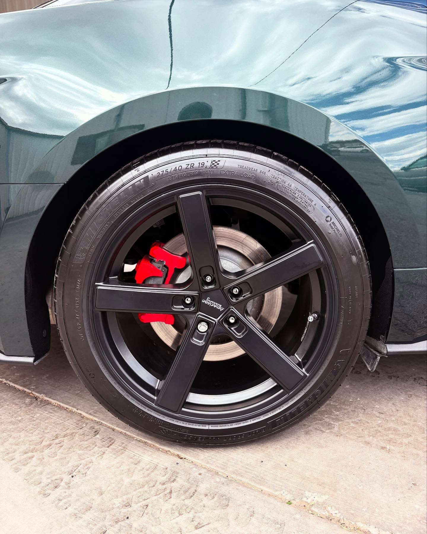 Close-up of a dark green car's rear wheel with a matte black five-spoke rim and a bright red brake caliper.