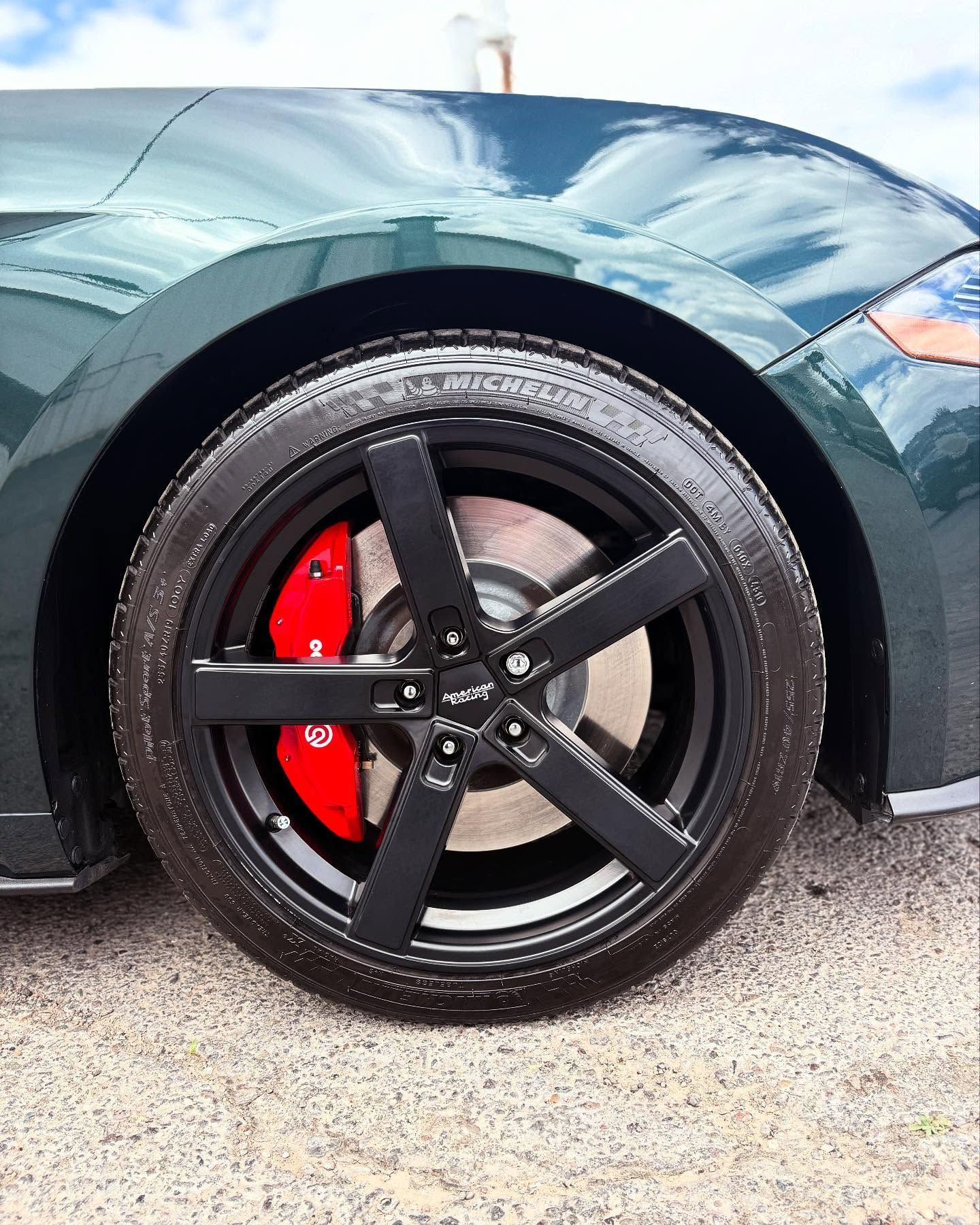 Close-up of a green car's front wheel with a black five-spoke rim and a prominent red brake caliper.