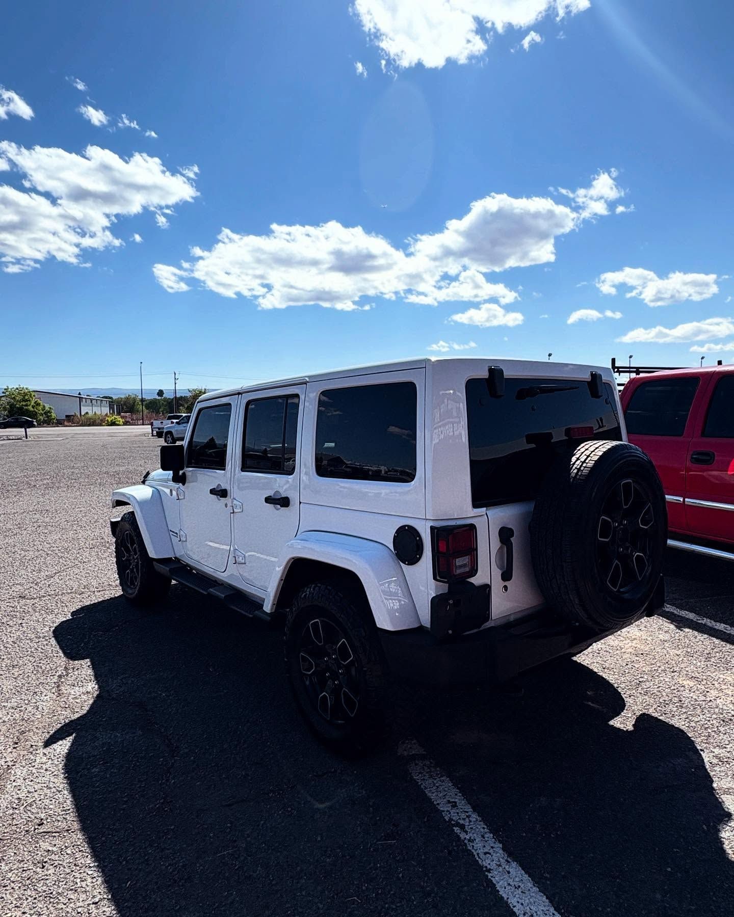 A white four-door Jeep Wrangler parked on a gravel lot under a clear, sunny blue sky with scattered clouds.